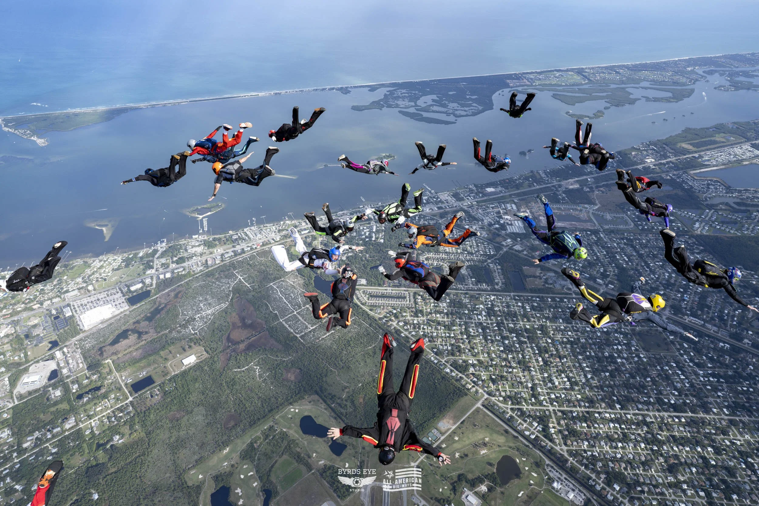 Group of skydivers in freefall over a cityscape with water bodies and green areas visible below.
