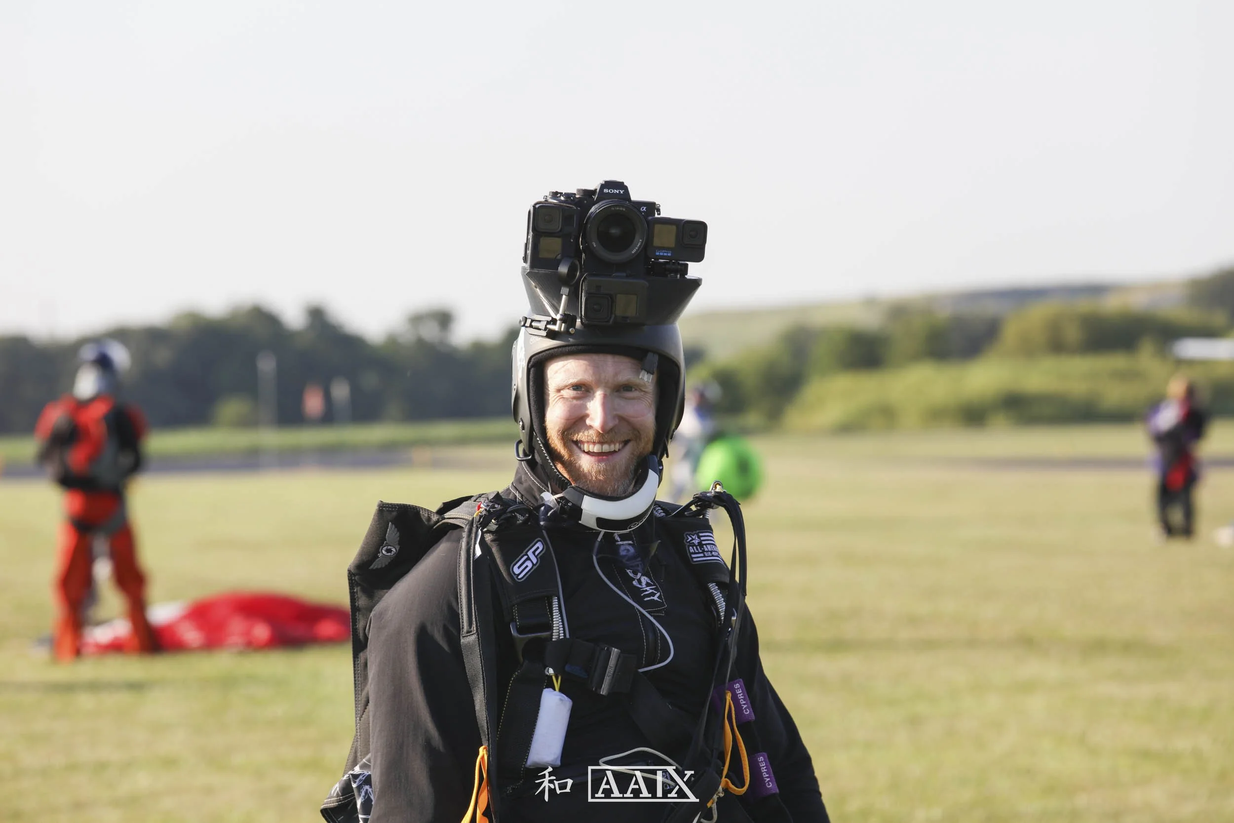 A man in skydiving gear smiling with a camera mounted on his helmet, standing on a grassy field with other skydivers in the background.