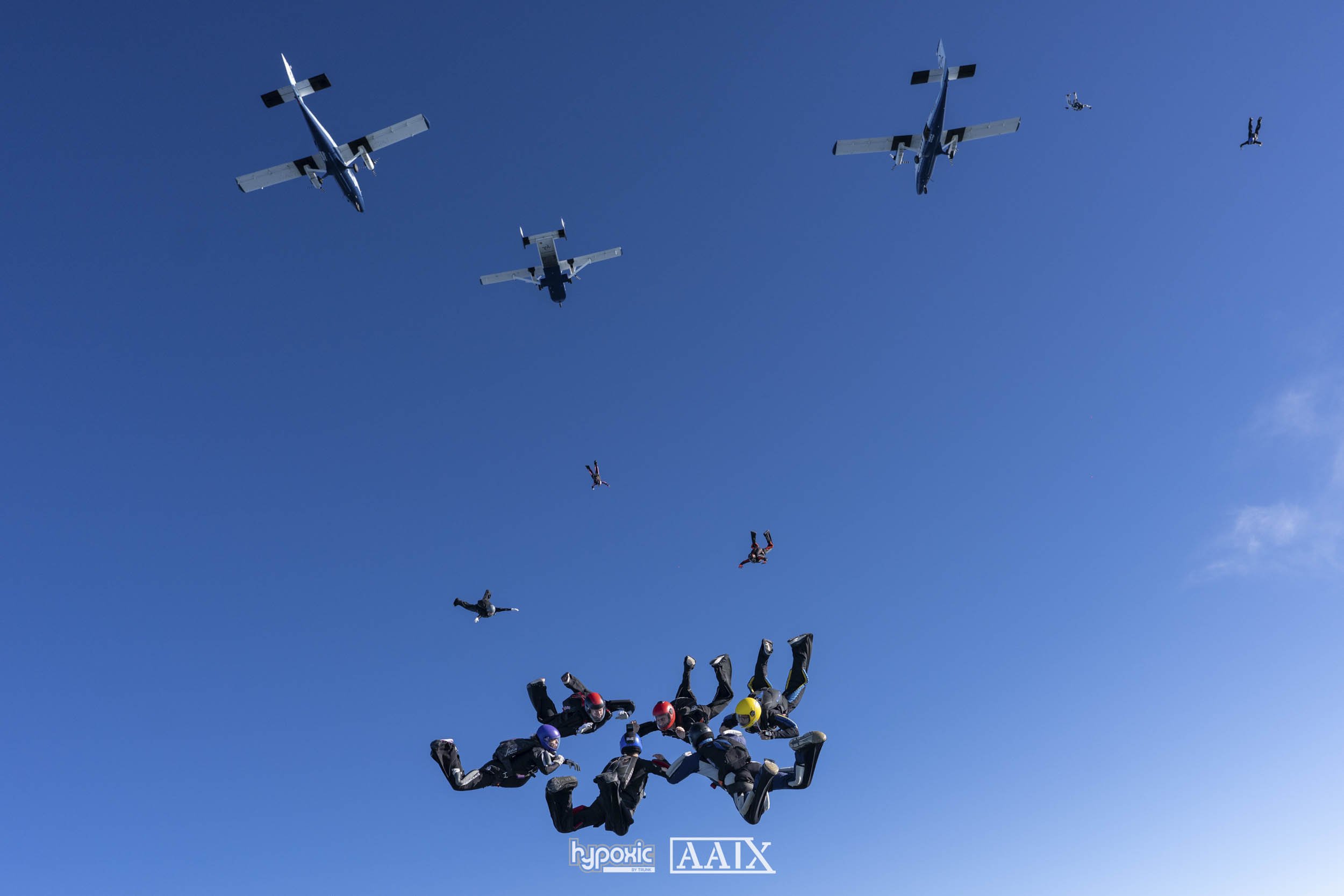 Skydivers in free fall formation with airplanes flying above against a clear blue sky.