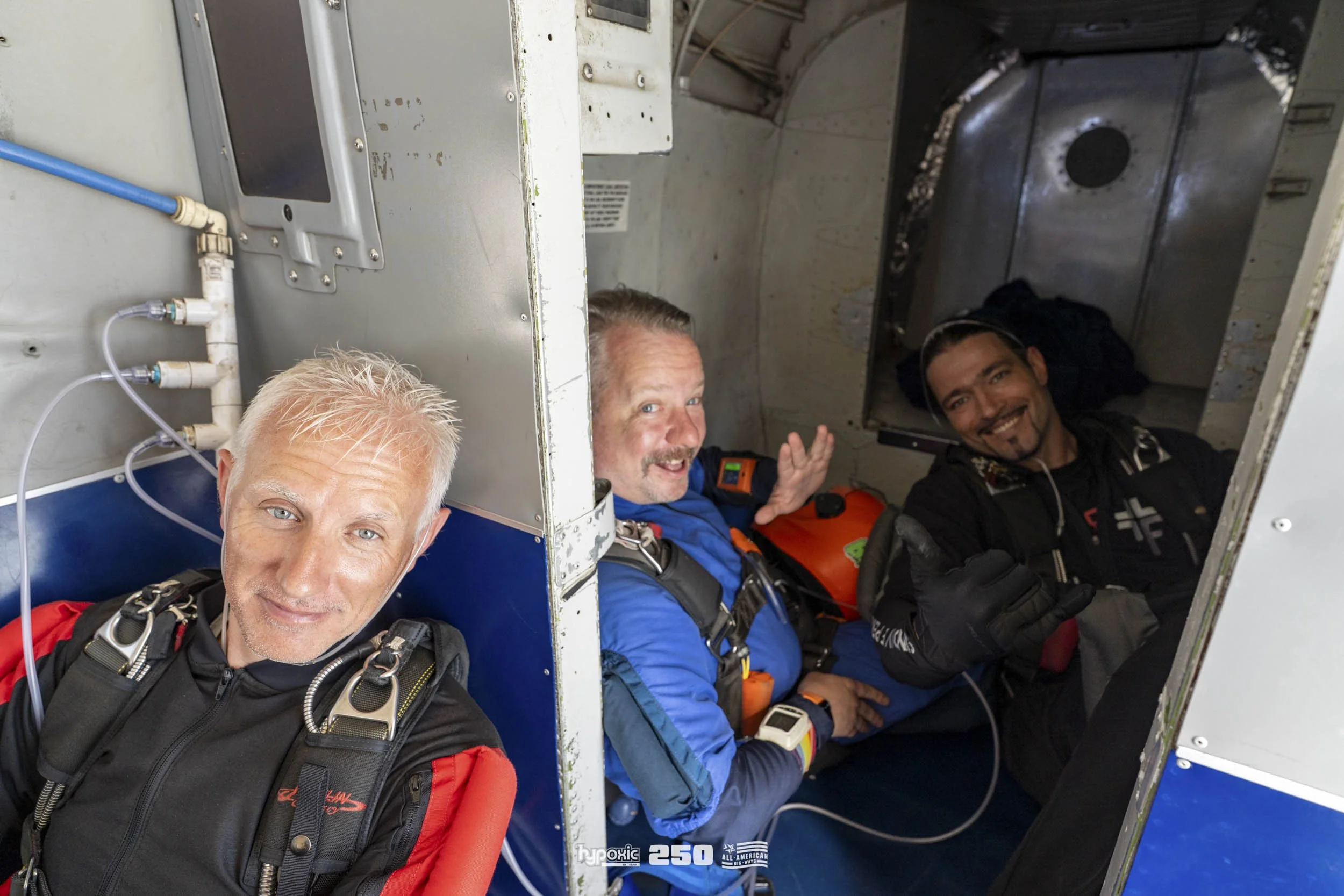 Three skydivers inside a parachute aircraft, seated and smiling before a jump.