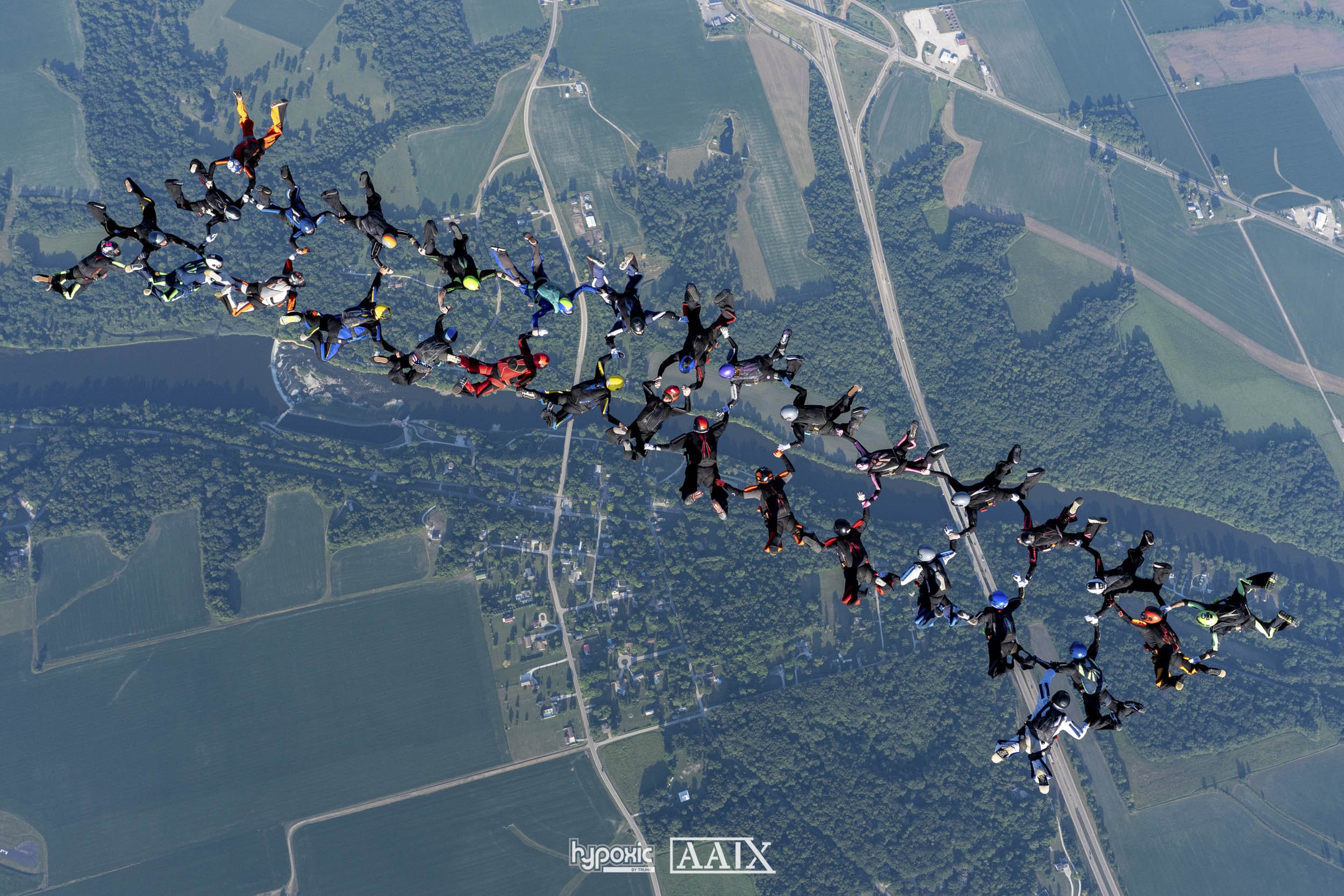 A group of skydivers holding hands forming a large chain in mid-air above a rural landscape with fields, roads, and trees.