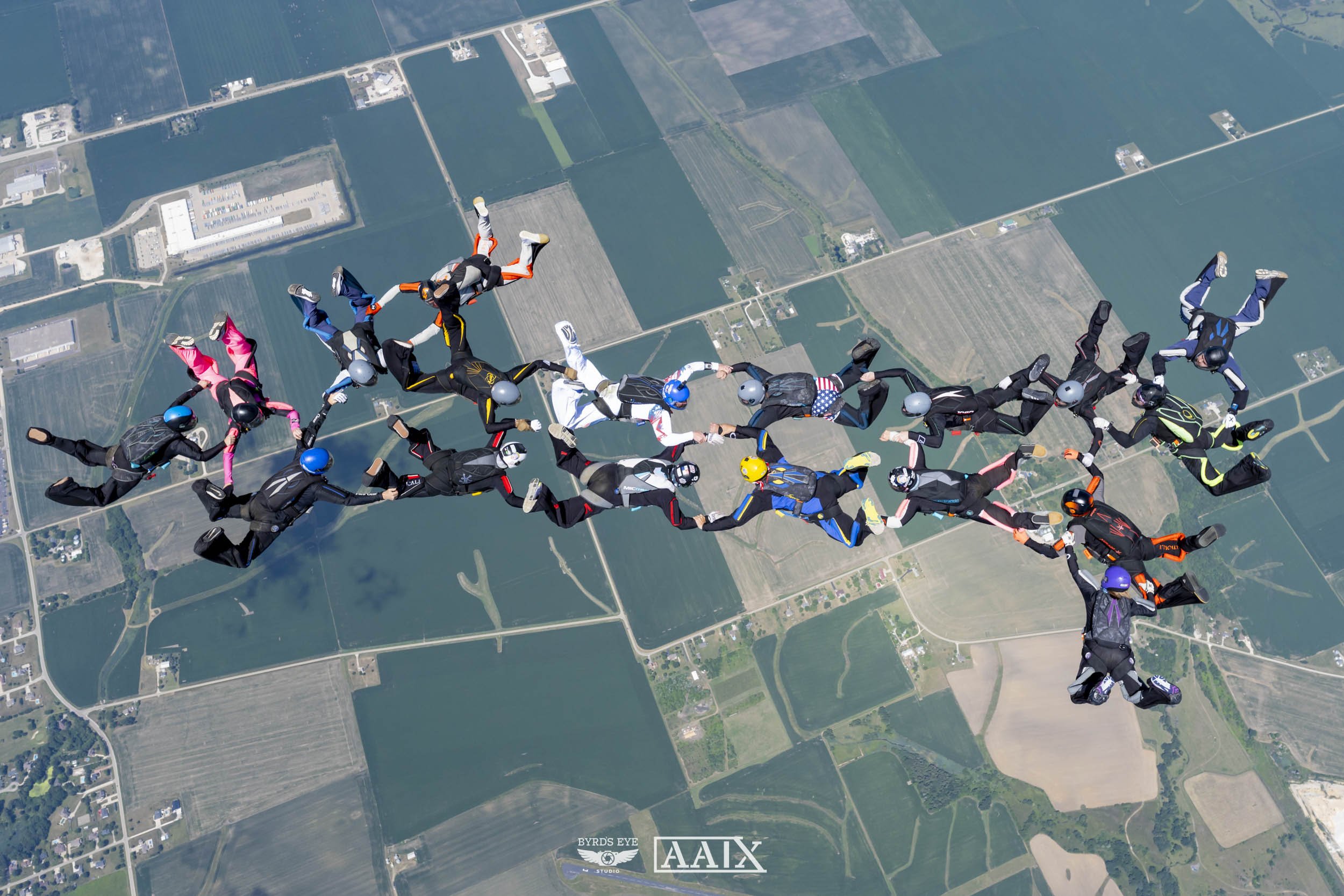 A group of skydivers holding hands in a formation while free falling over a landscape of farmland and water bodies.