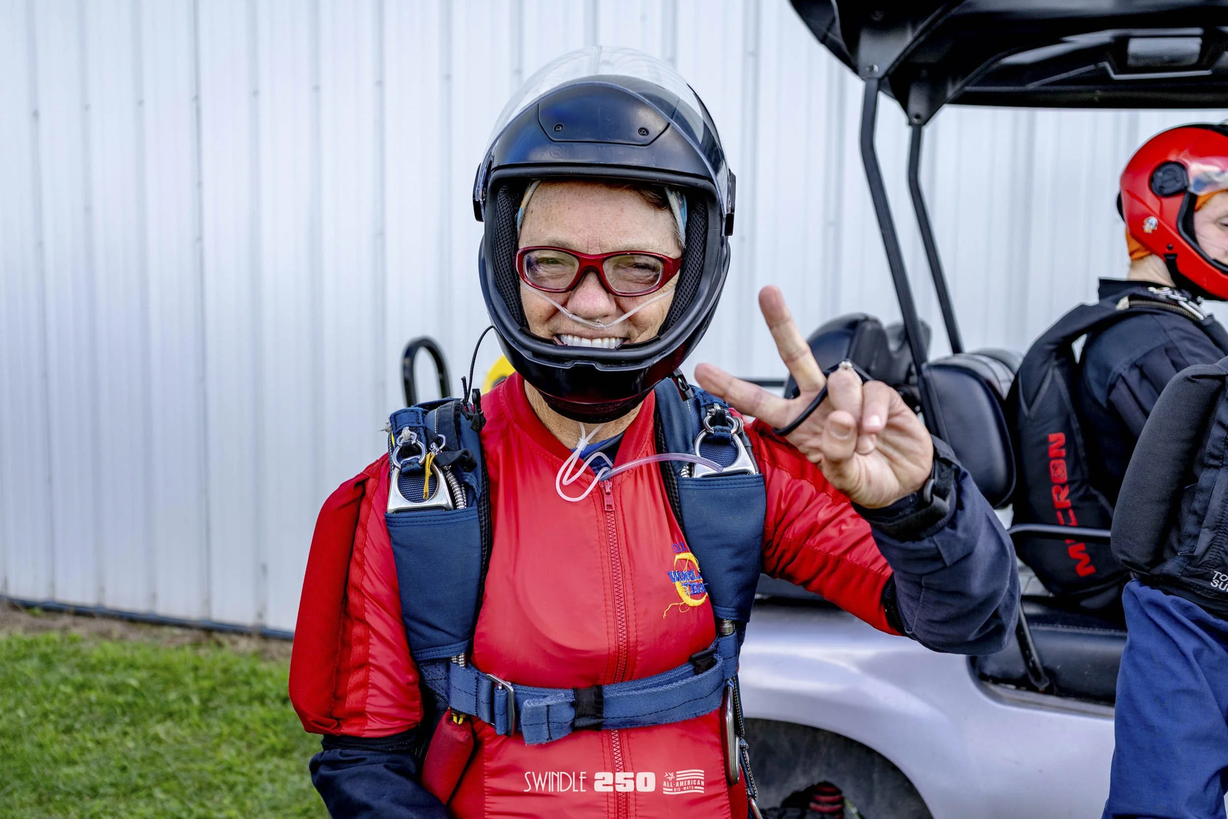 A smiling woman in a red skydiving jumpsuit and helmet making a peace sign outdoors next to a small aircraft.