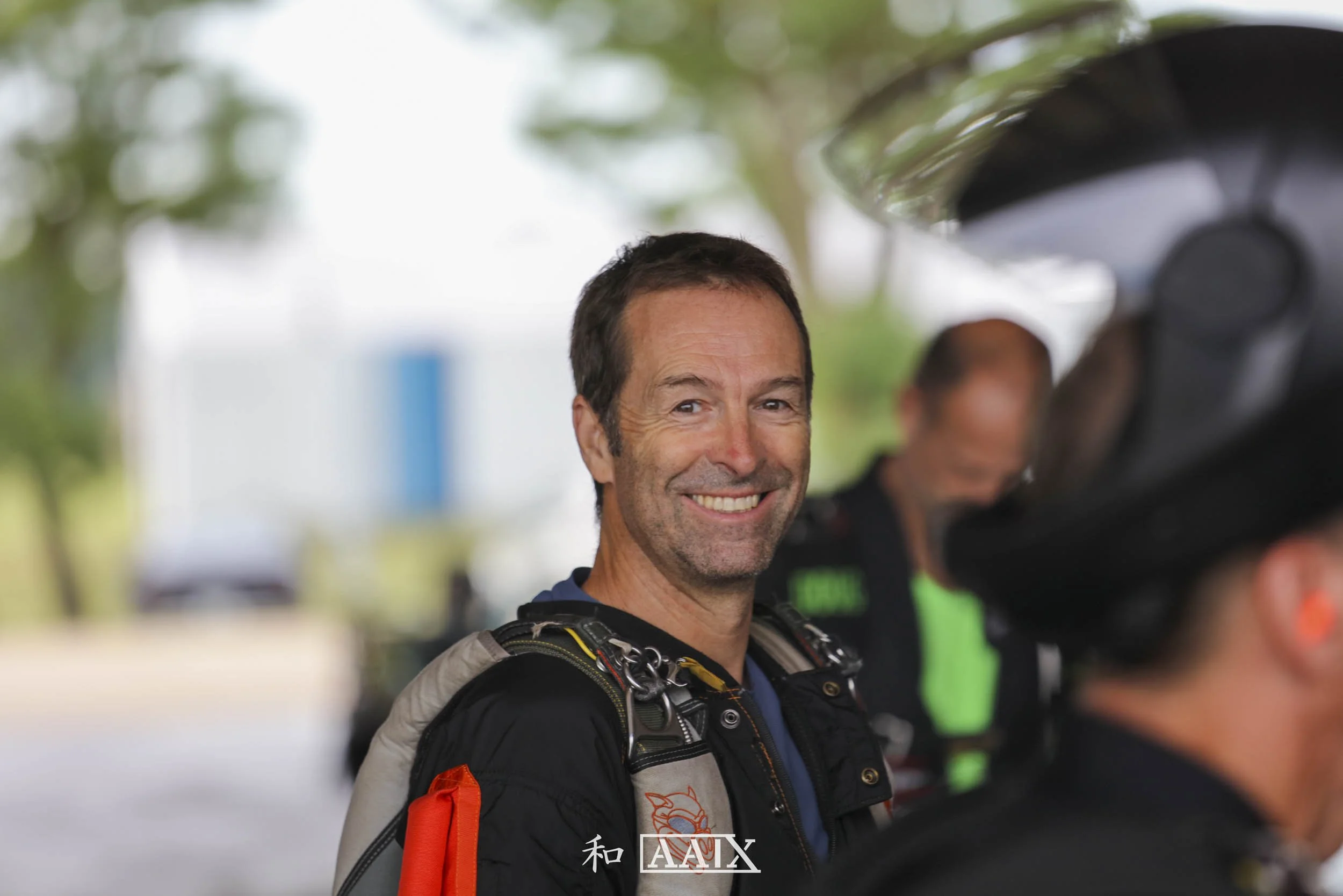 A smiling man in motorcycle gear standing outdoors with blurred trees and other people in the background.