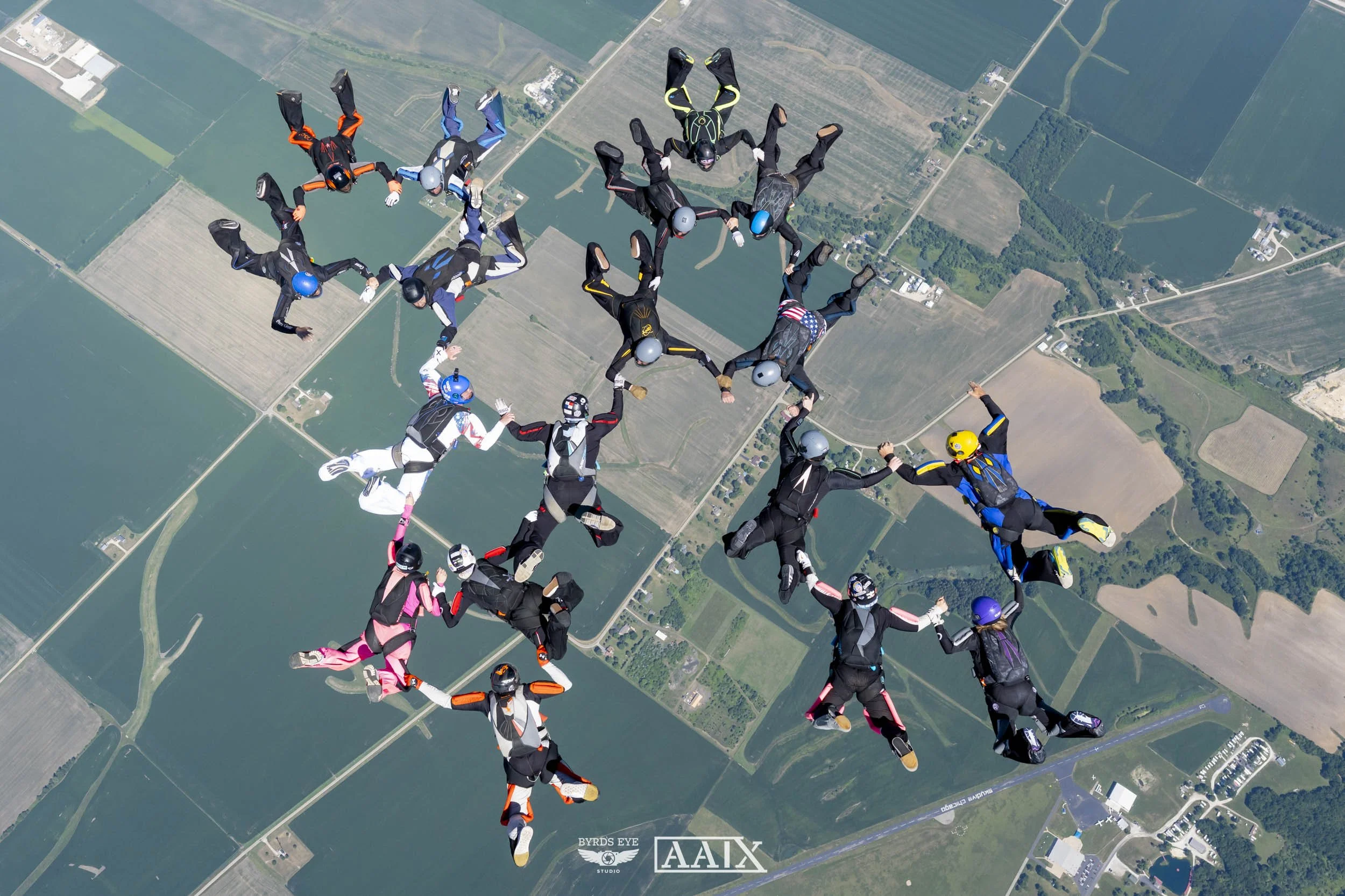 A large group of skydivers in colorful jumpsuits and helmets form a circle while mid-air over farmland and fields, holding hands in a coordinated formation.