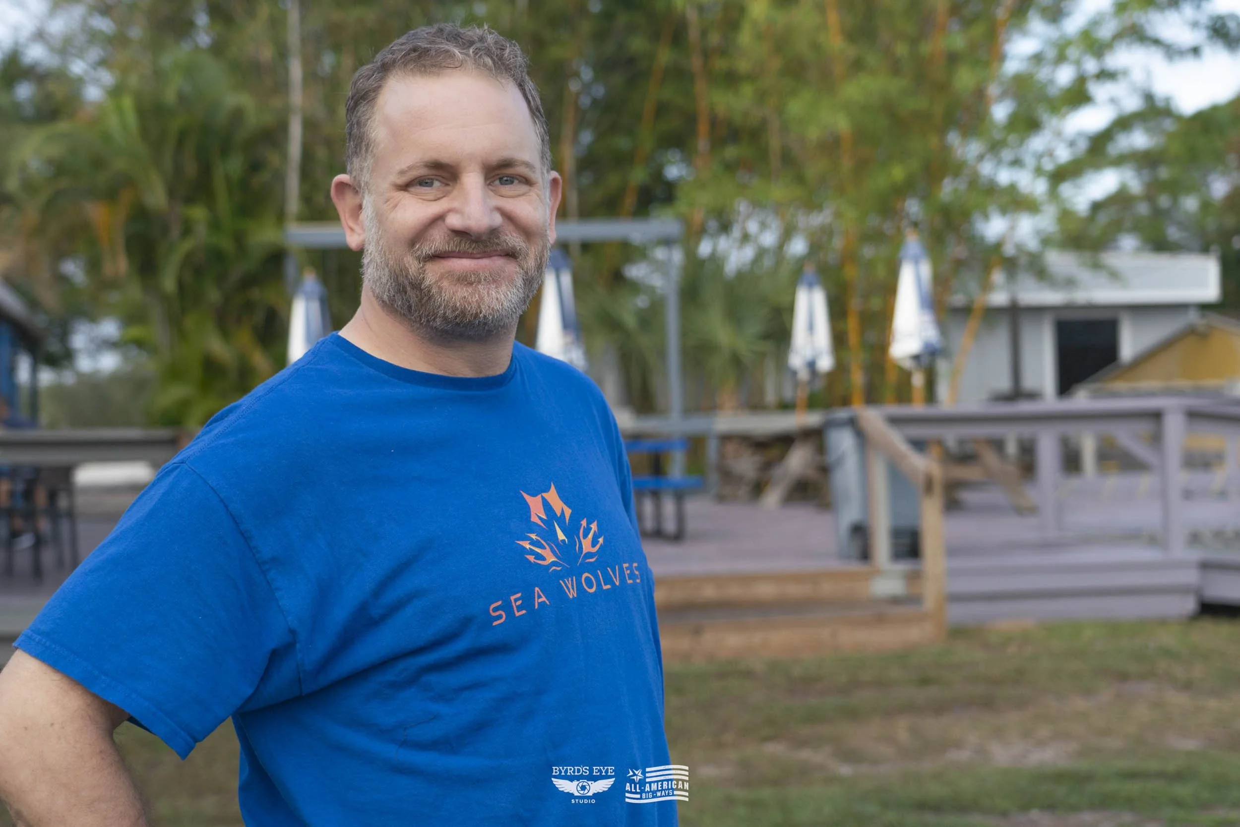 Smiling man with a beard wearing a blue t-shirt with a logo and text, standing outdoors near a deck and trees.