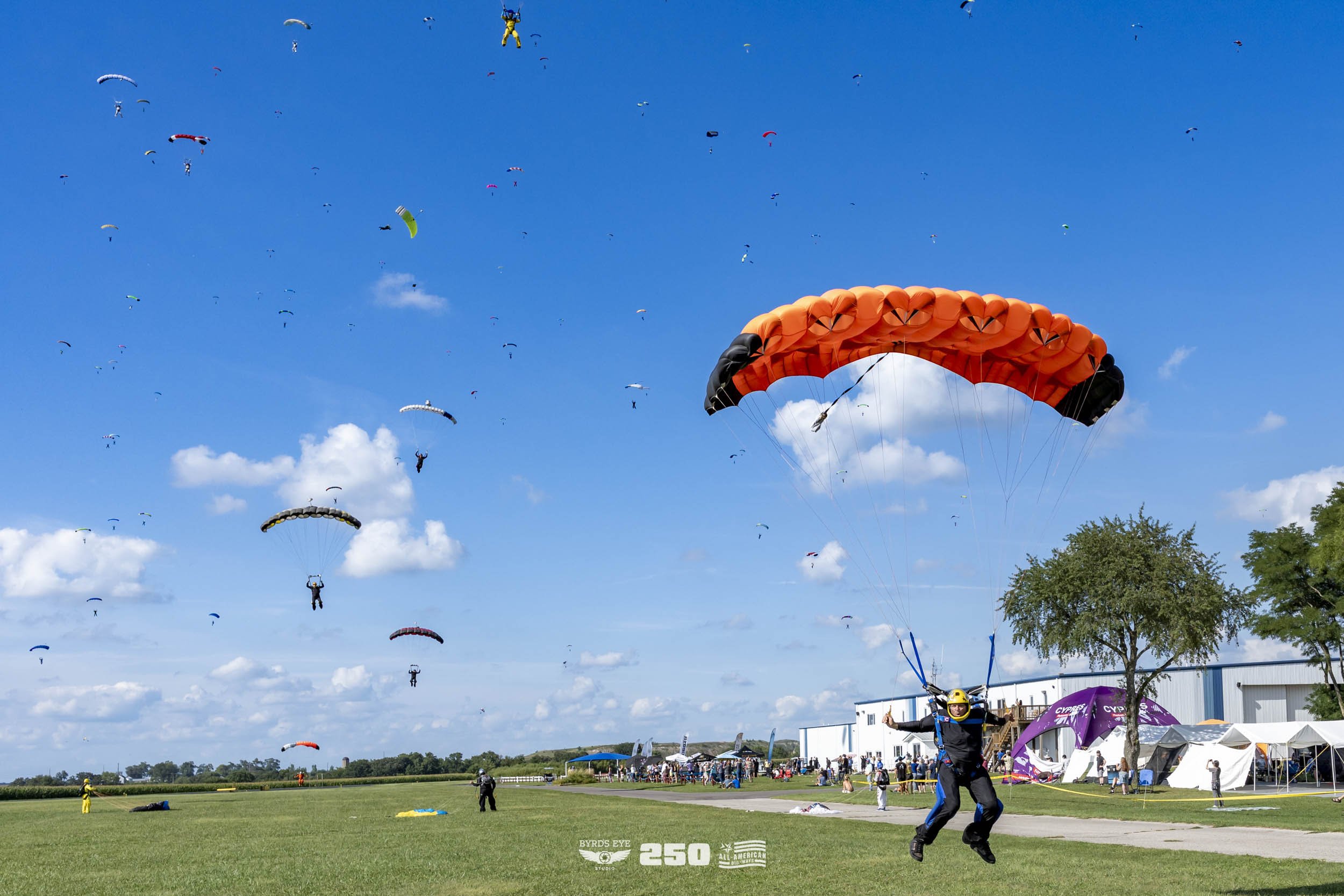 A person with a yellow helmet and black outfit parachuting with an orange and black parachute at a skydiving event, with many other parachutes in the blue sky and a crowd of spectators and tents on the ground.