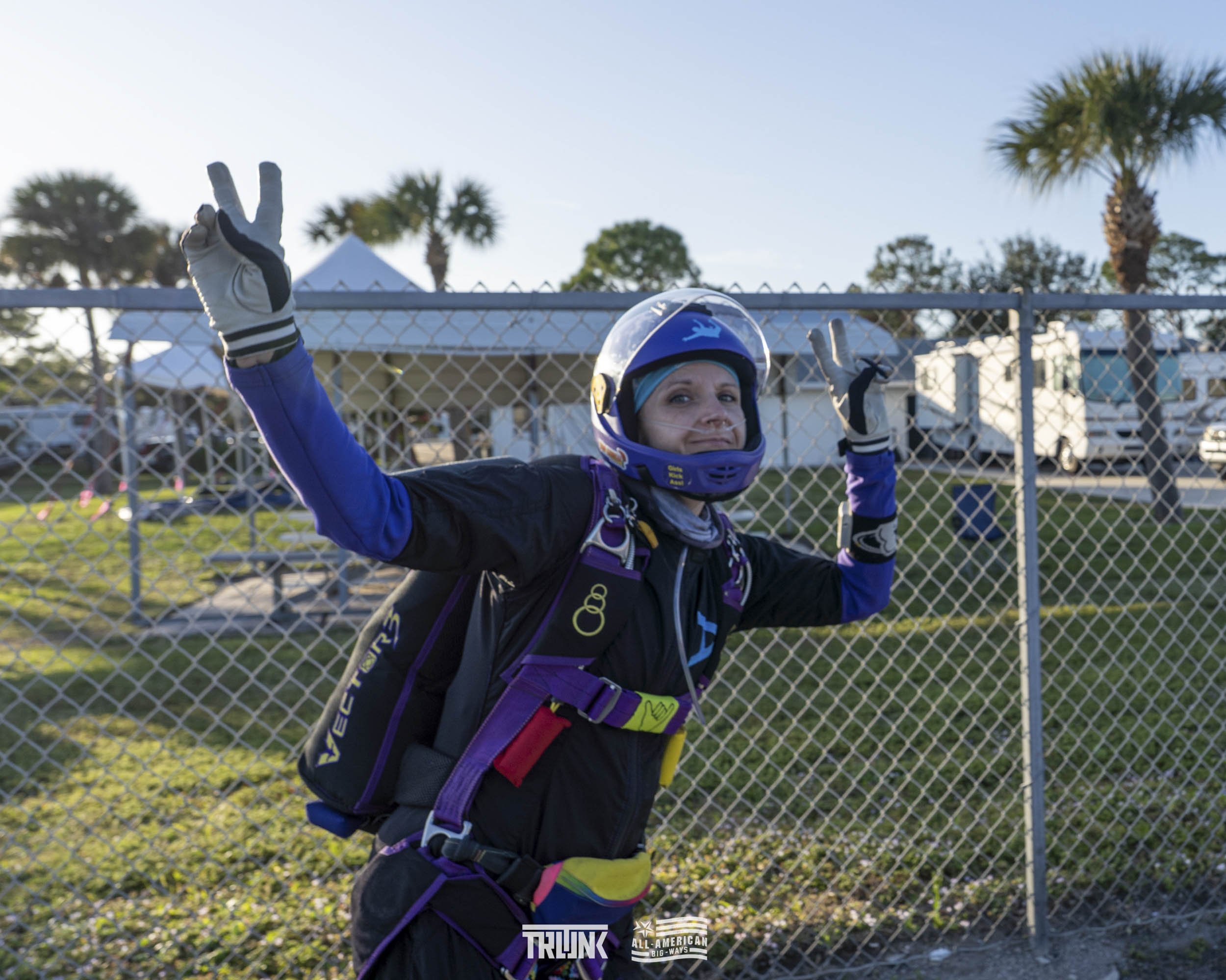 A woman in skydiving gear making a peace sign with both hands in front of a chain-link fence with trees and parked RVs in the background.