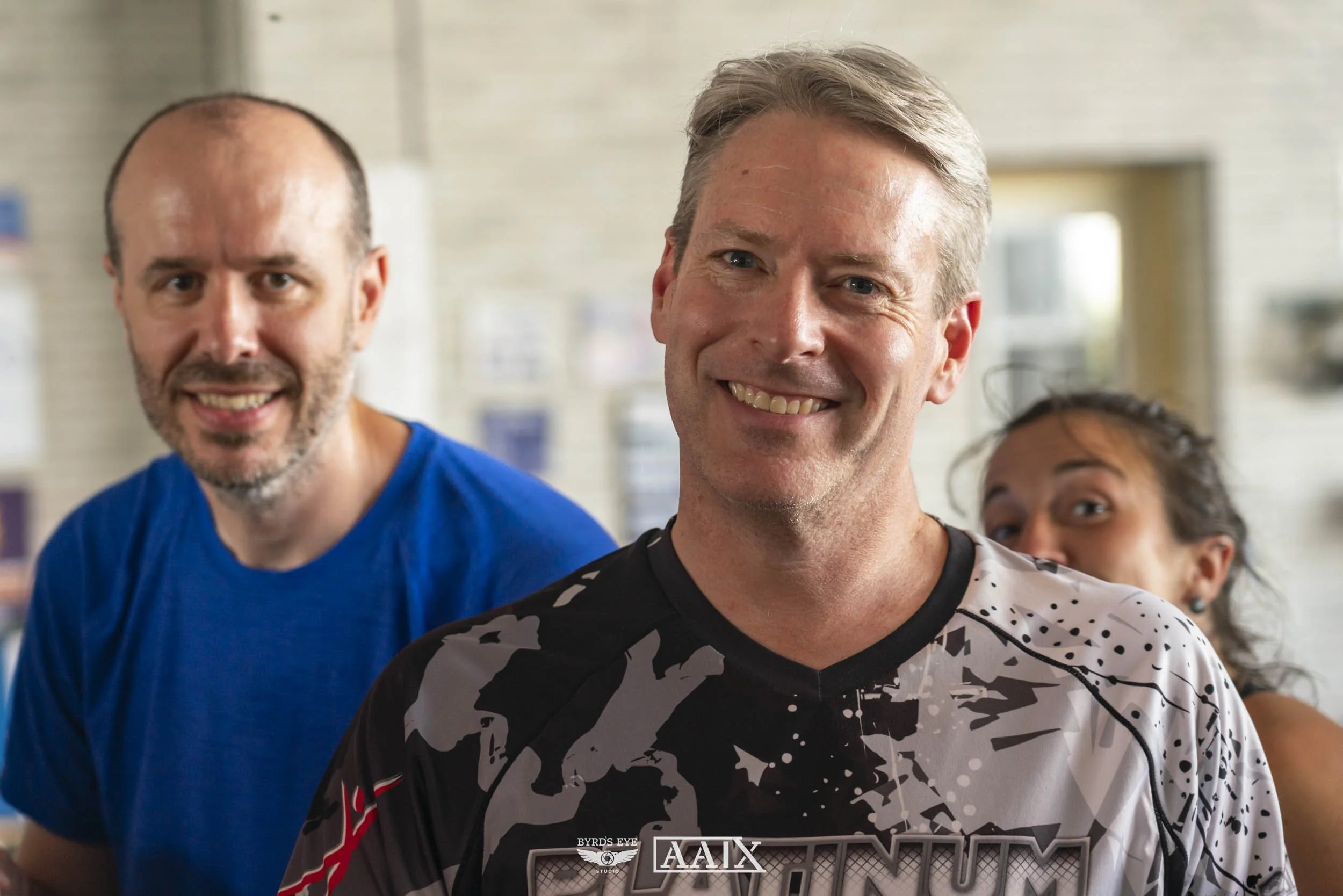 Three smiling people, two men and one woman, standing indoors in front of a wall with pictures or notices. The man in the foreground is wearing a black and white sports shirt, the man behind him is wearing a blue shirt, and the woman peeking from beh