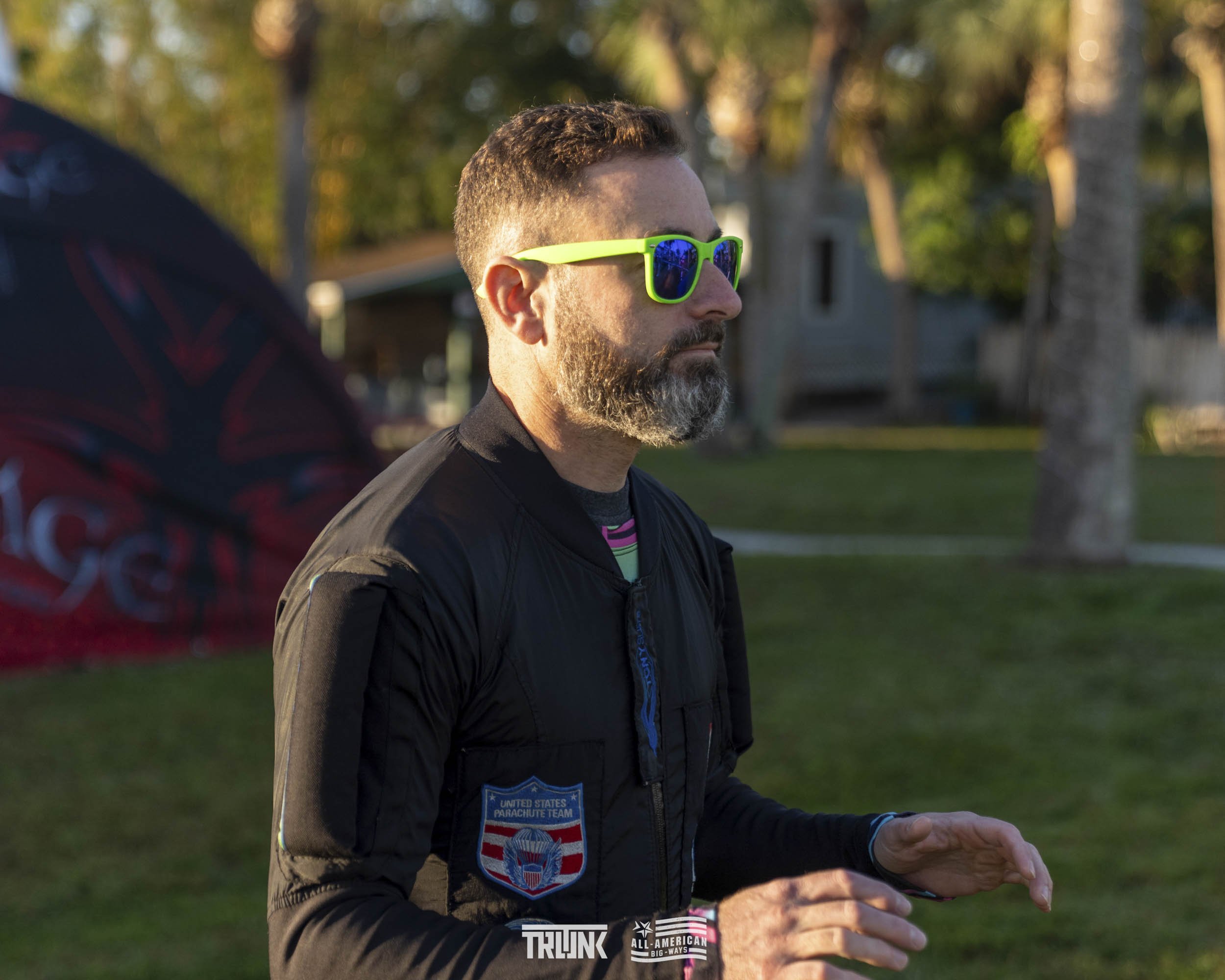 A middle-aged man with a beard and sunglasses standing outdoors in a park or grassy area during the daytime.
