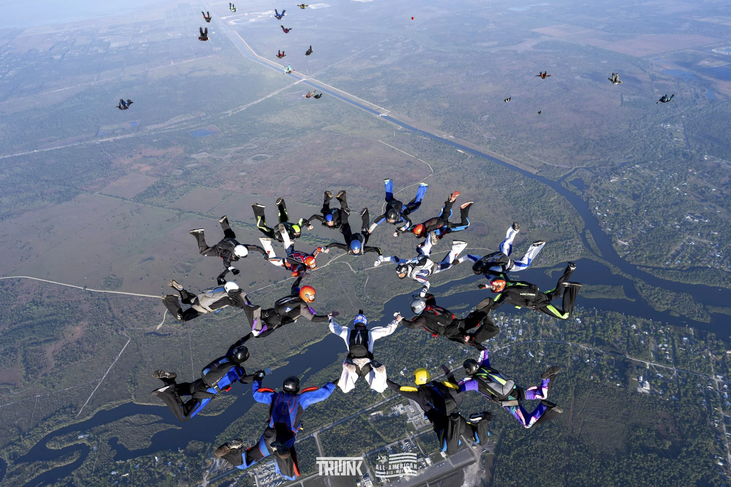 A large group of skydivers holding hands forms a star shape while free-falling above a landscape with rivers and farmland.