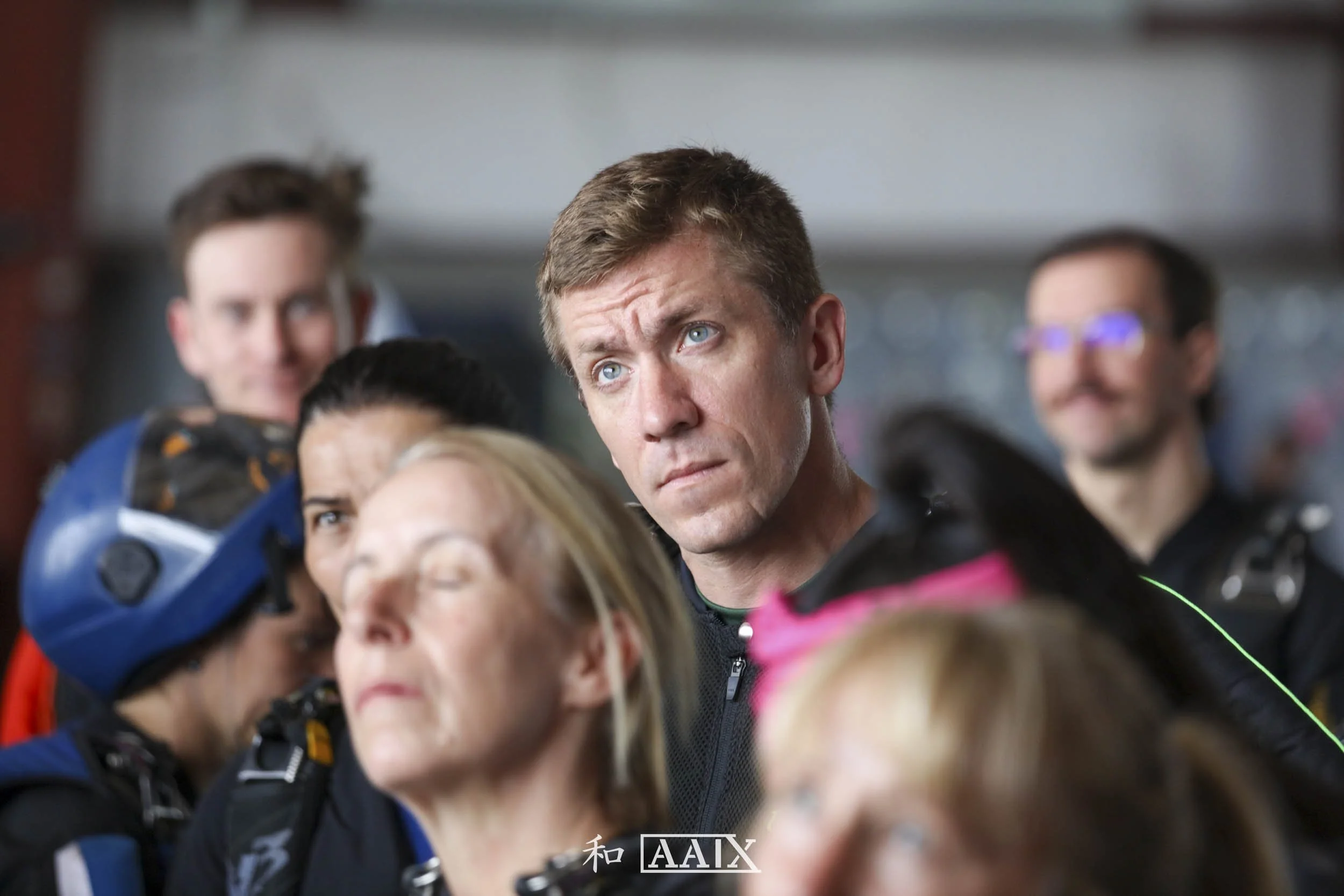A man with short light brown hair and blue eyes looks intently to the side in a group of people, some wearing helmets and outdoor gear, in what appears to be an indoor setting.