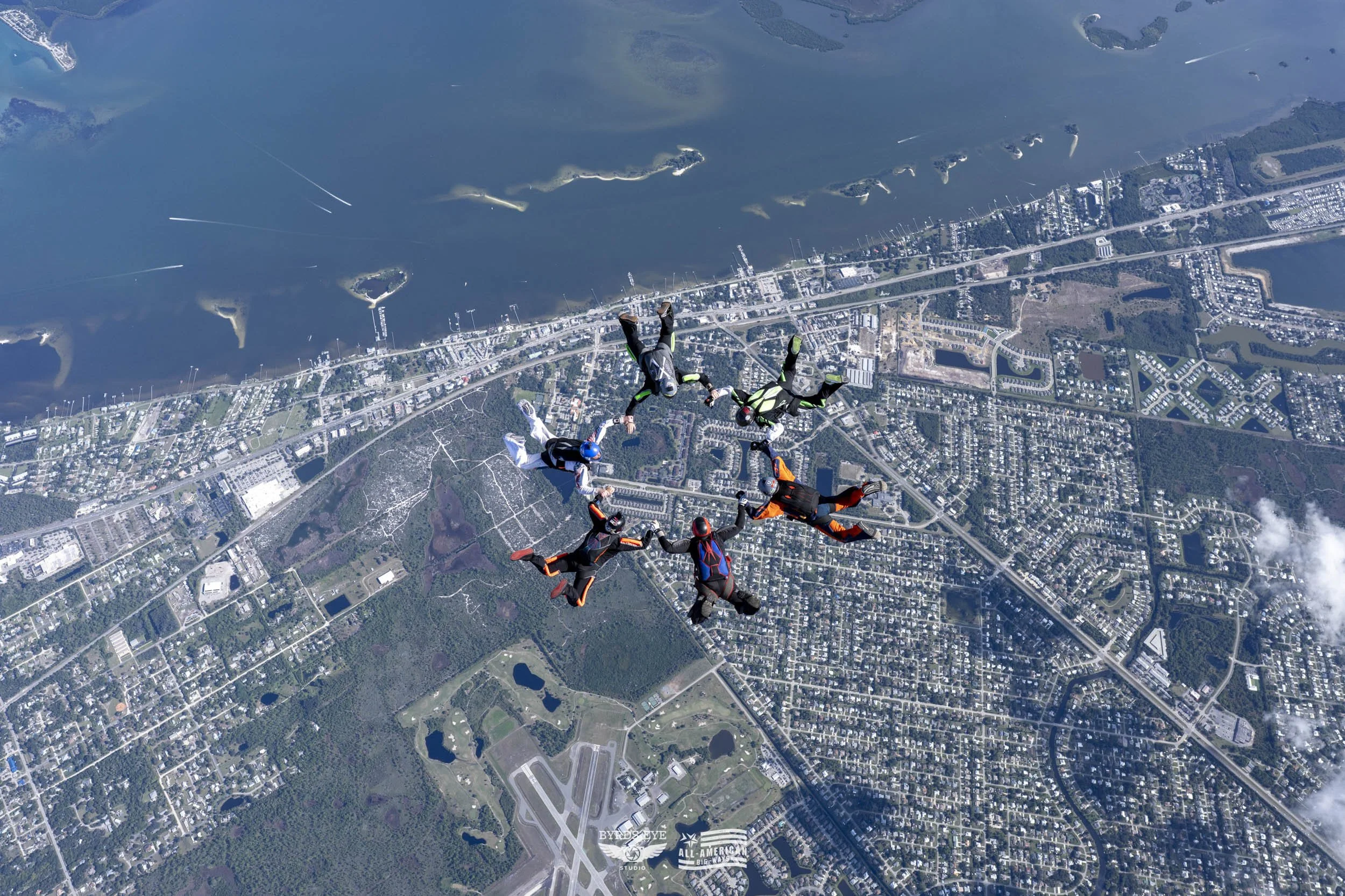A group of skydivers holding hands in a circle free-falling over a city and body of water.