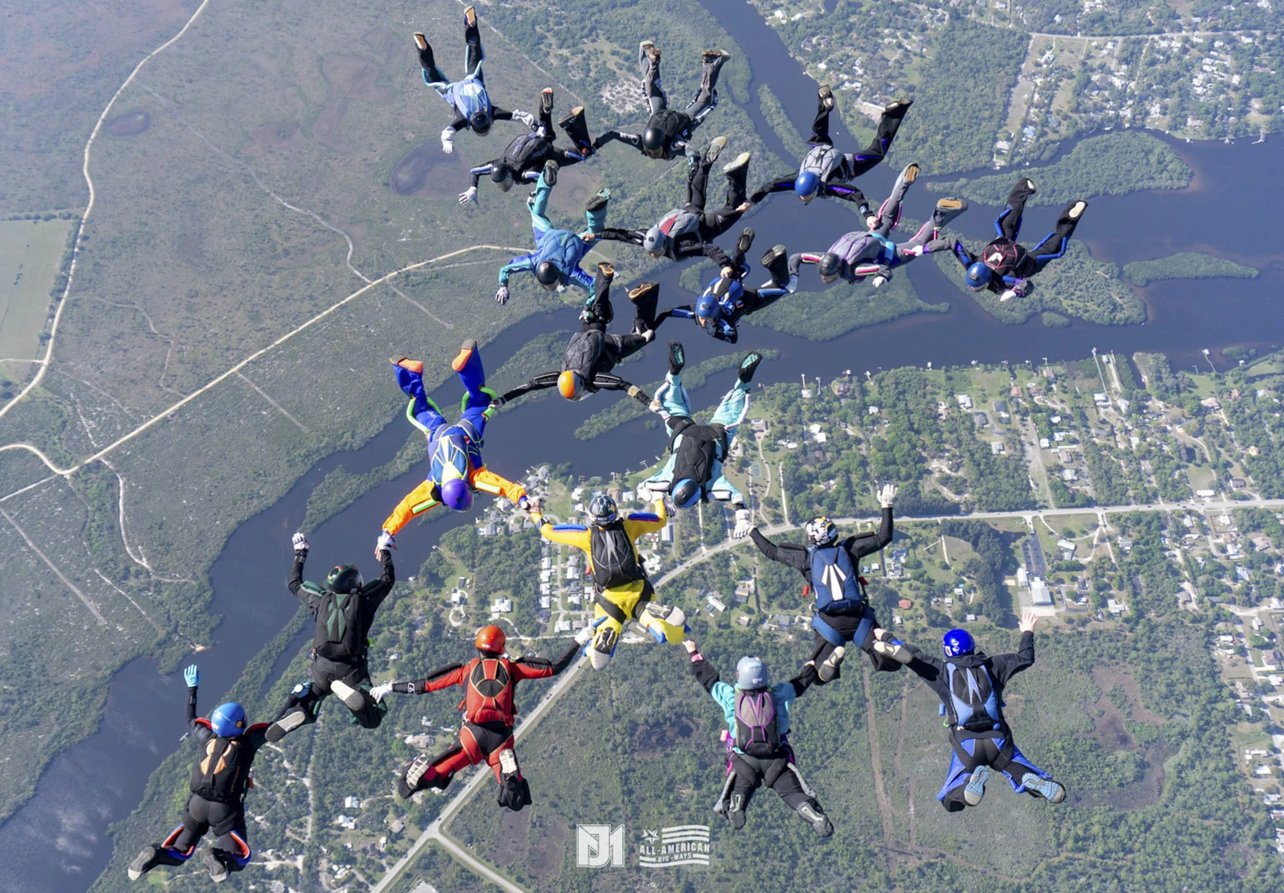 Group of skydivers in colorful suits and helmets holding hands in a formation during a jump over a landscape with lakes, forests, and roads.
