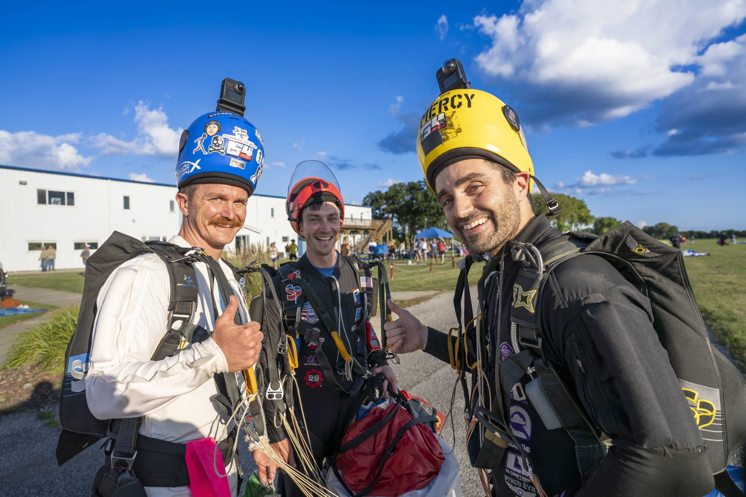 Three men in skydiving gear smiling outside, with parachutes and helmets, on a sunny day with a blue sky and clouds, in front of a white building and open field.