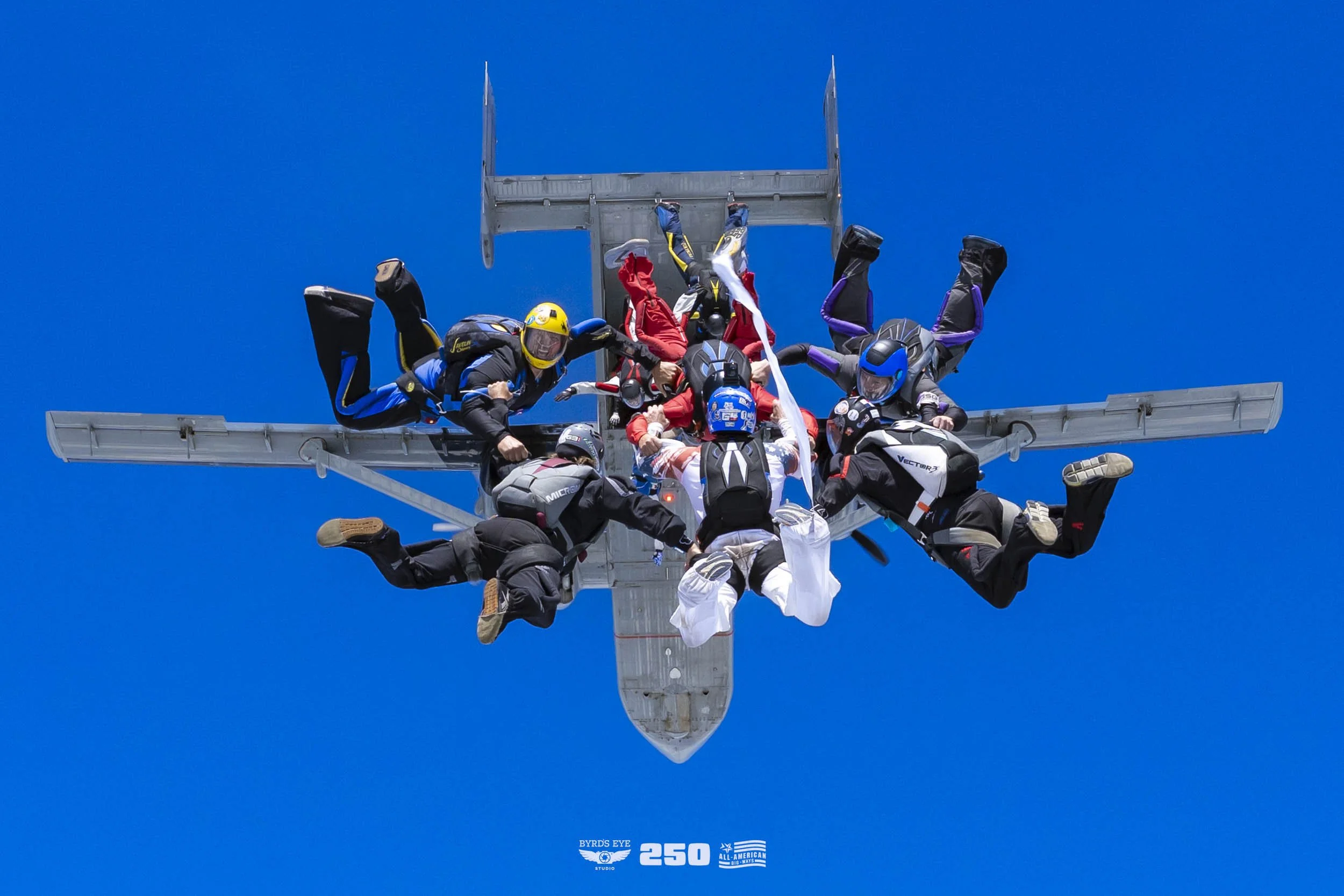 Multiple skydivers in free fall beneath an airplane on a clear blue sky background, with some holding hands and others in different positions.