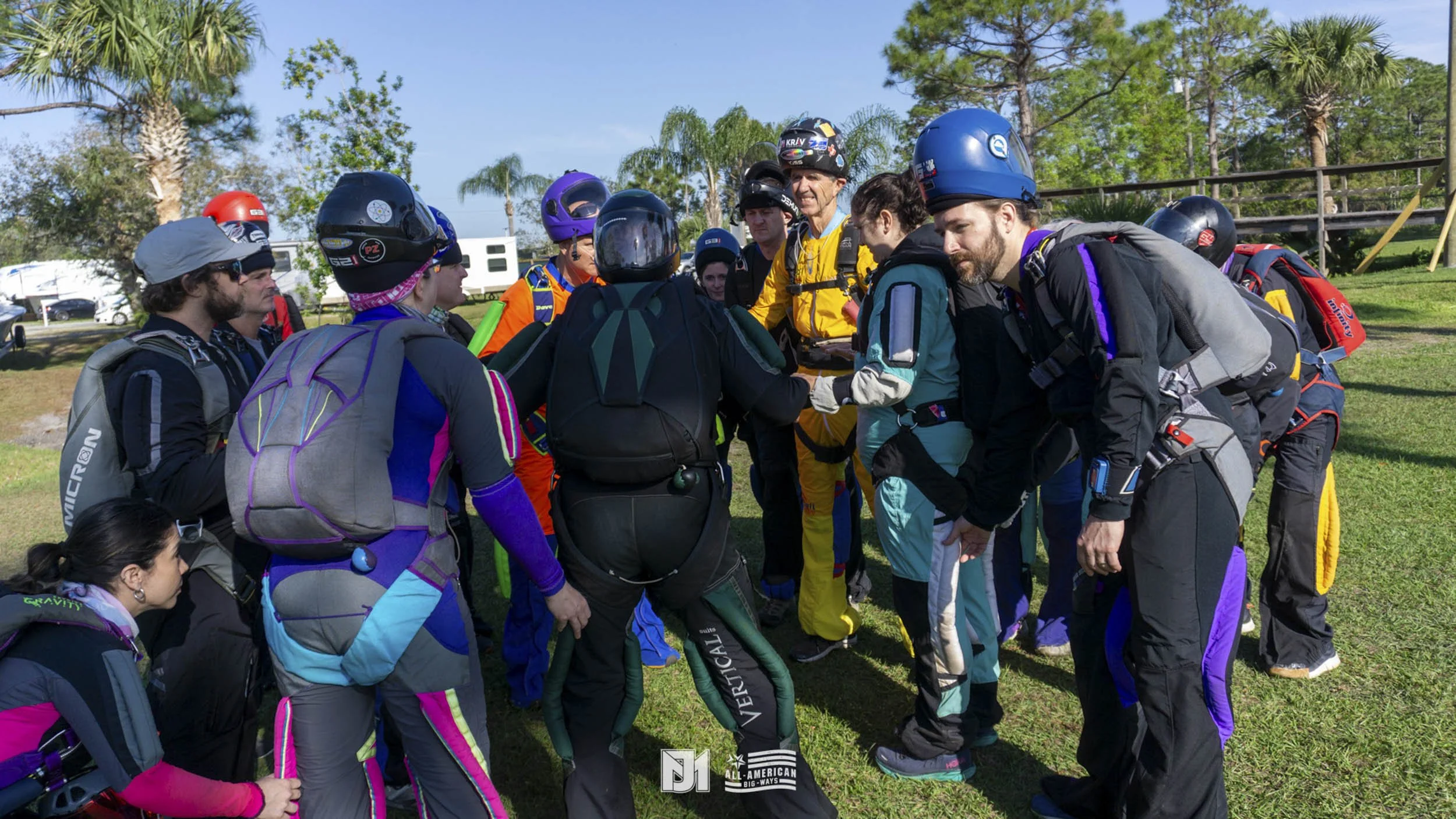 A group of skydivers in jumpsuits and helmets gathered in a circle outdoors on a grassy field, preparing for a jump.