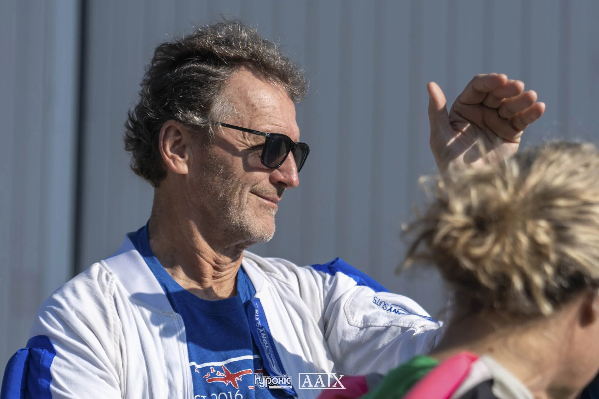 A man with dark sunglasses and gray hair, wearing a blue and white racing jacket, smiling and raising his hand towards a woman with curly blonde hair, who is partially visible. They are outdoors against a gray building.