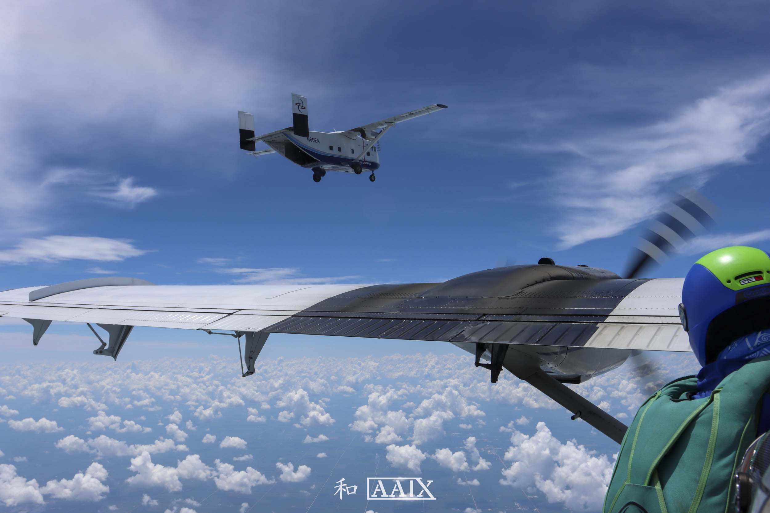 A person in skydiving gear, including a helmet and harness, is in a wingsuit flying through the sky below a small aircraft, with another plane flying above against a backdrop of blue sky and clouds.