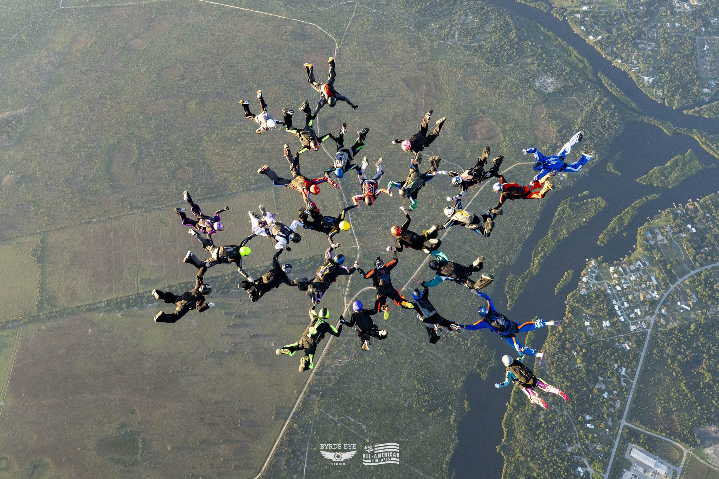 Group of skydivers in colorful jumpsuits and helmets forming a star shape while free-falling over a landscape with lakes and fields.