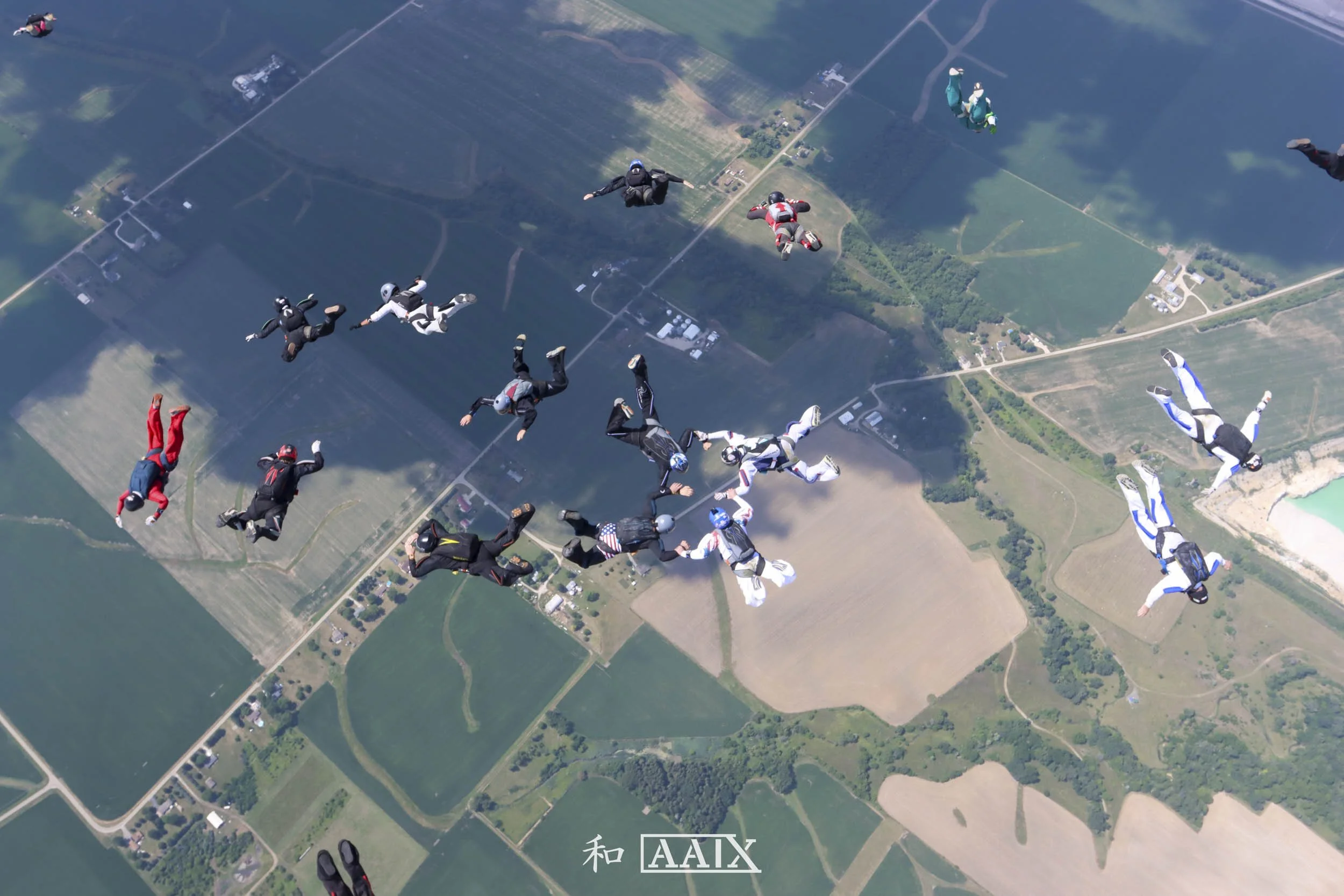 Multiple skydivers in freefall over a patchwork of farmland fields, some holding hands in a circle.