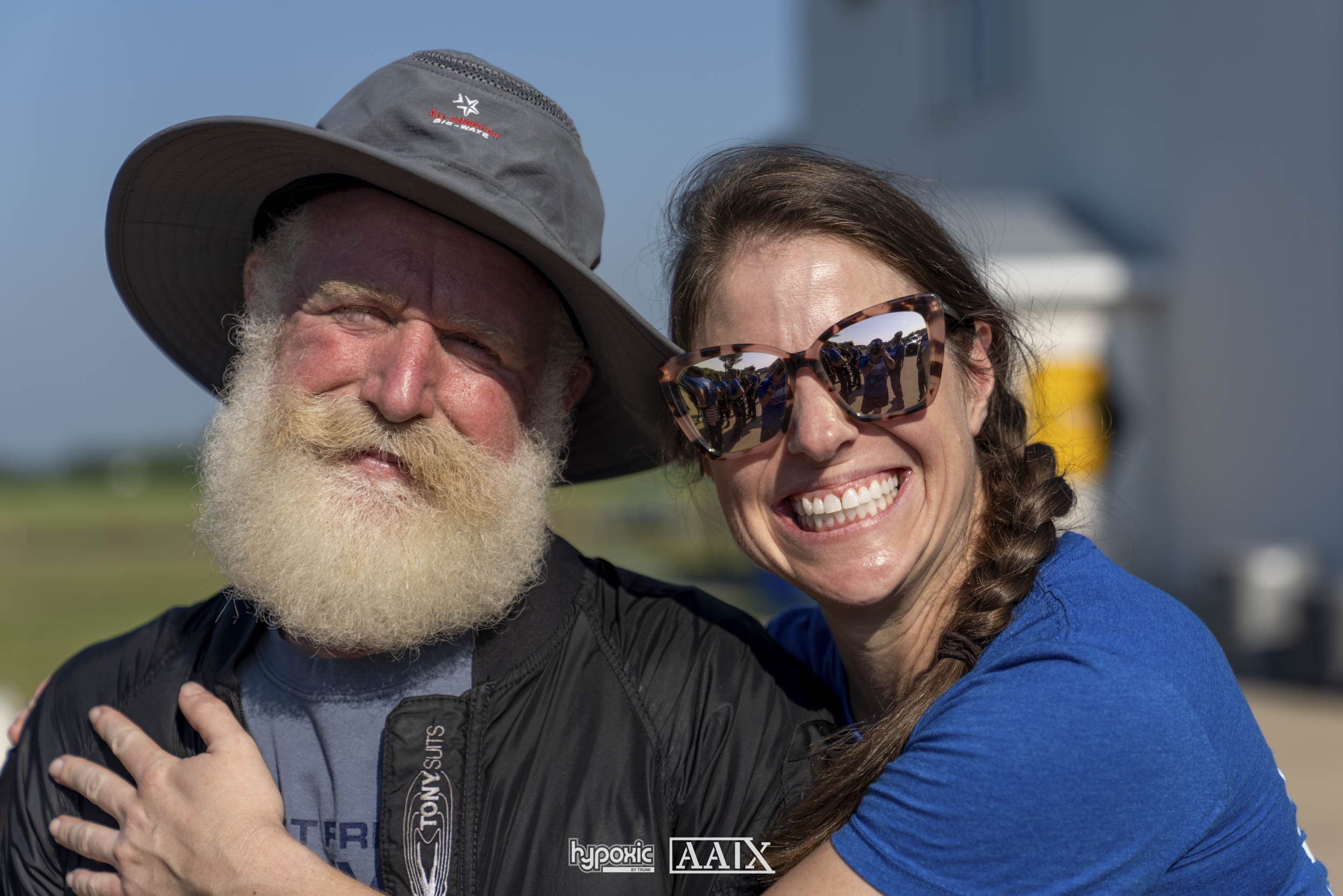 A cheerful man with a large gray sun hat and a white beard is hugging a woman with long braided hair, sunglasses, and a bright smile outdoors on a sunny day.