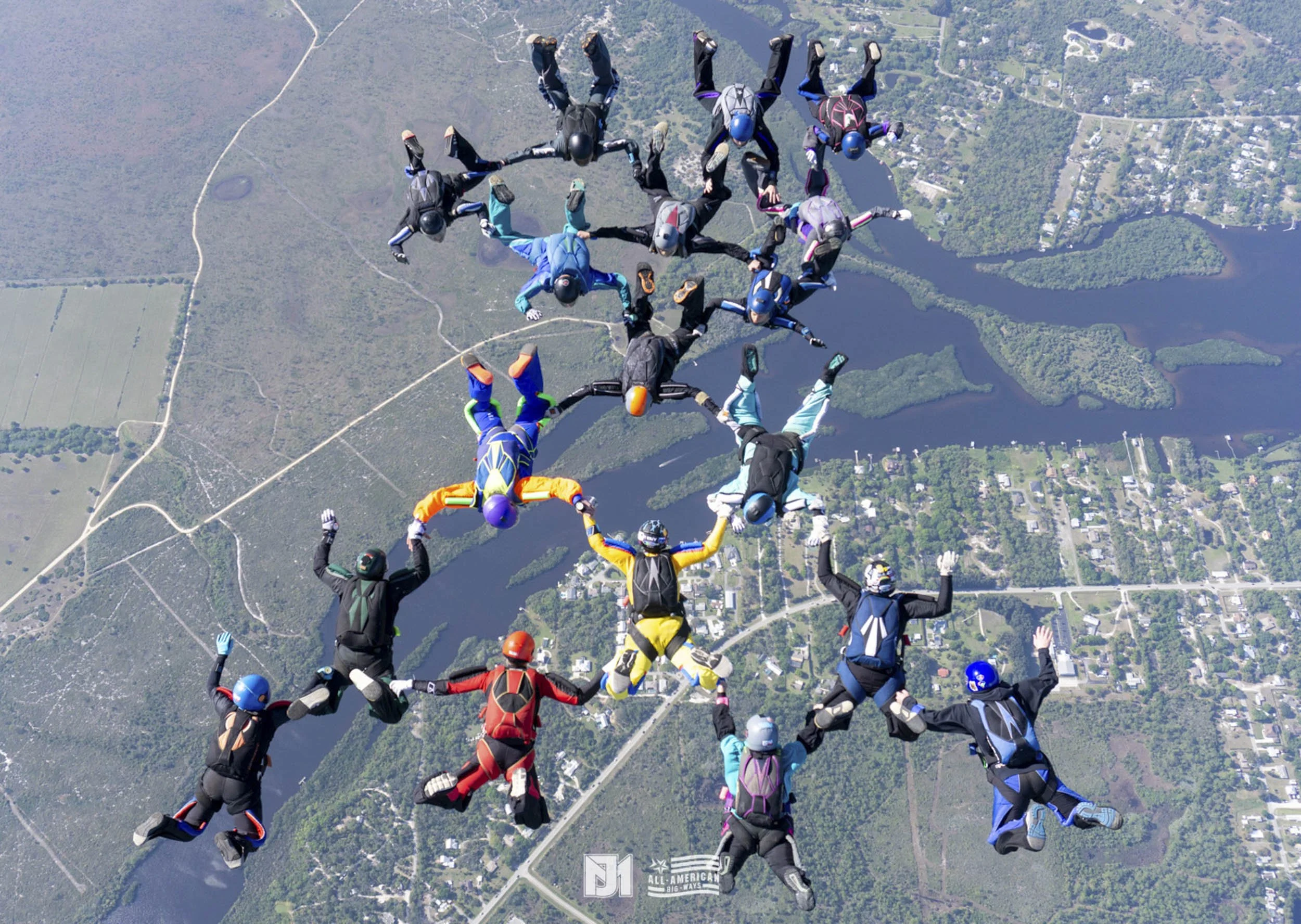 Group of skydivers in freefall formation over a landscape with lakes and roads.
