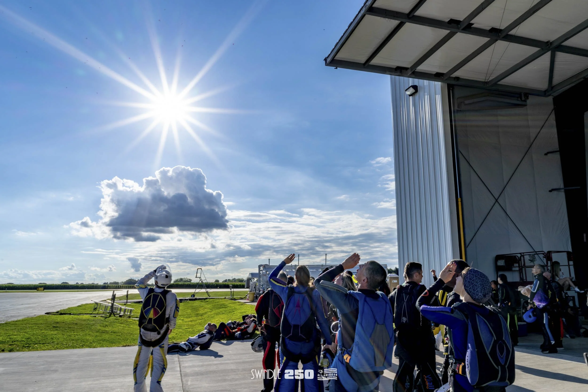 A group of skydivers on the ground at an airfield, preparing for a jump on a sunny day with a few clouds in the sky.