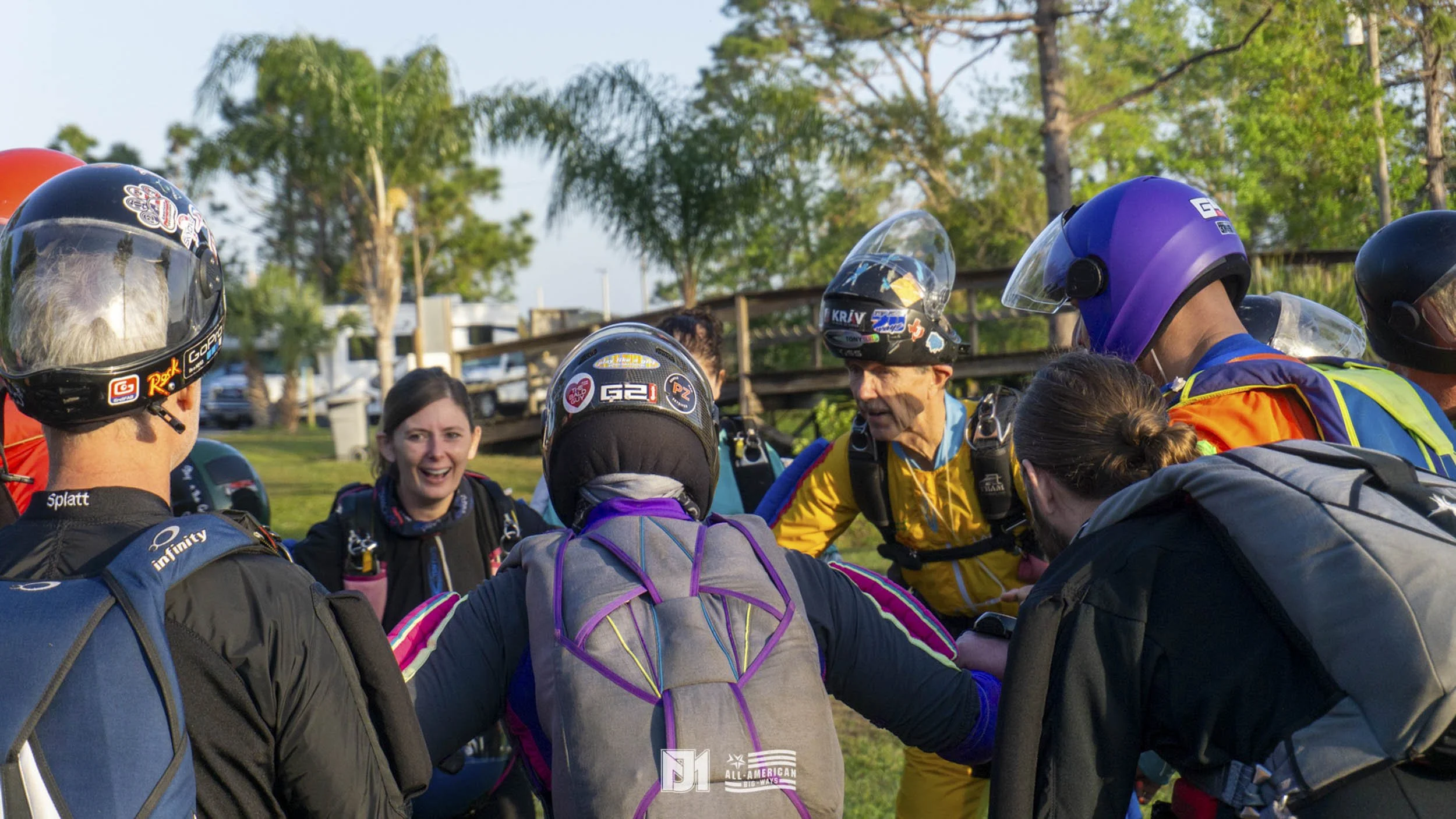 A group of skydivers wearing helmets and gear gathered in a circle outdoors, likely in preparation for a jump, with trees and a grassy area in the background.