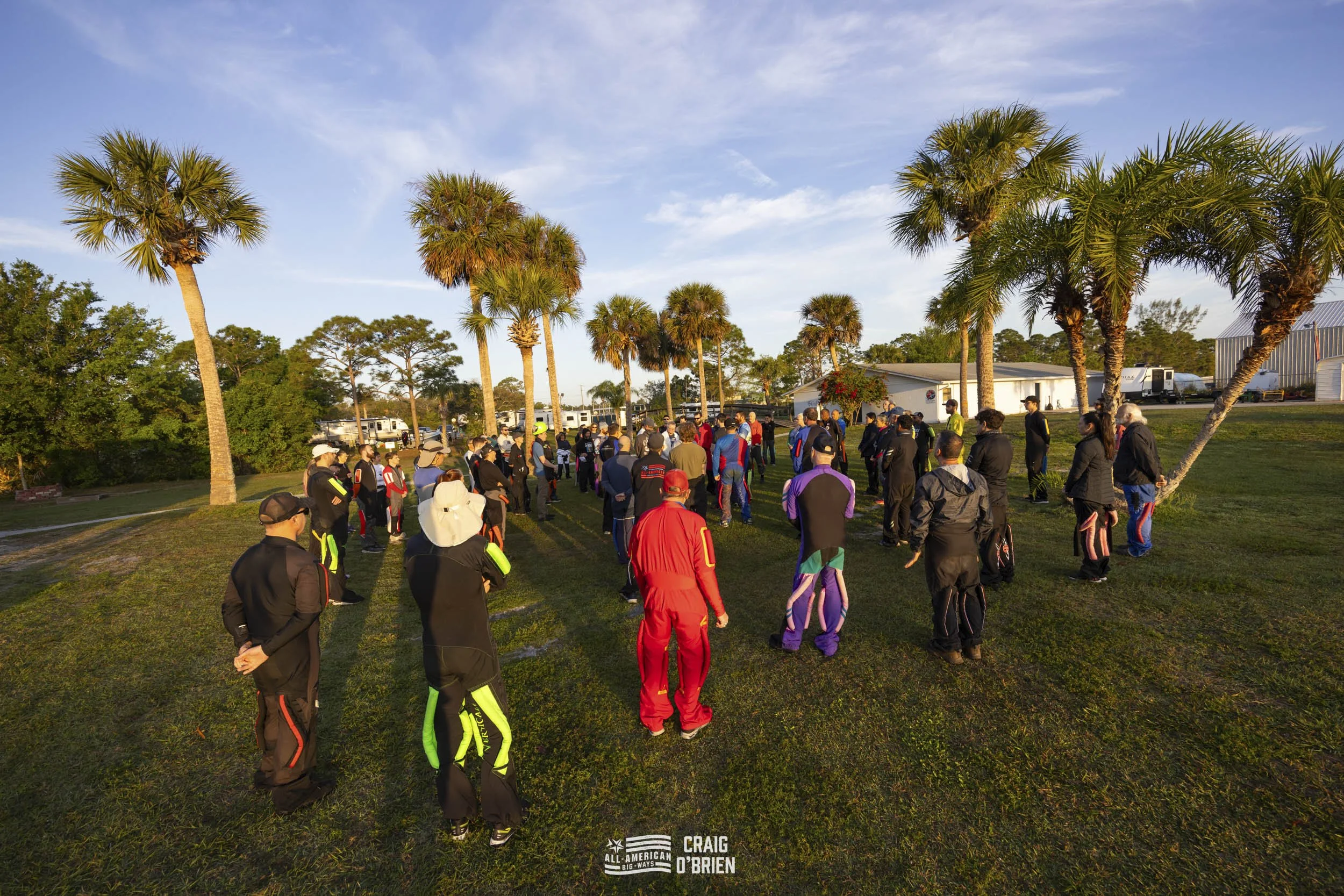 Group of people gathered outdoors on a grassy area with palm trees, possibly for an event or meeting, during late afternoon or early evening.