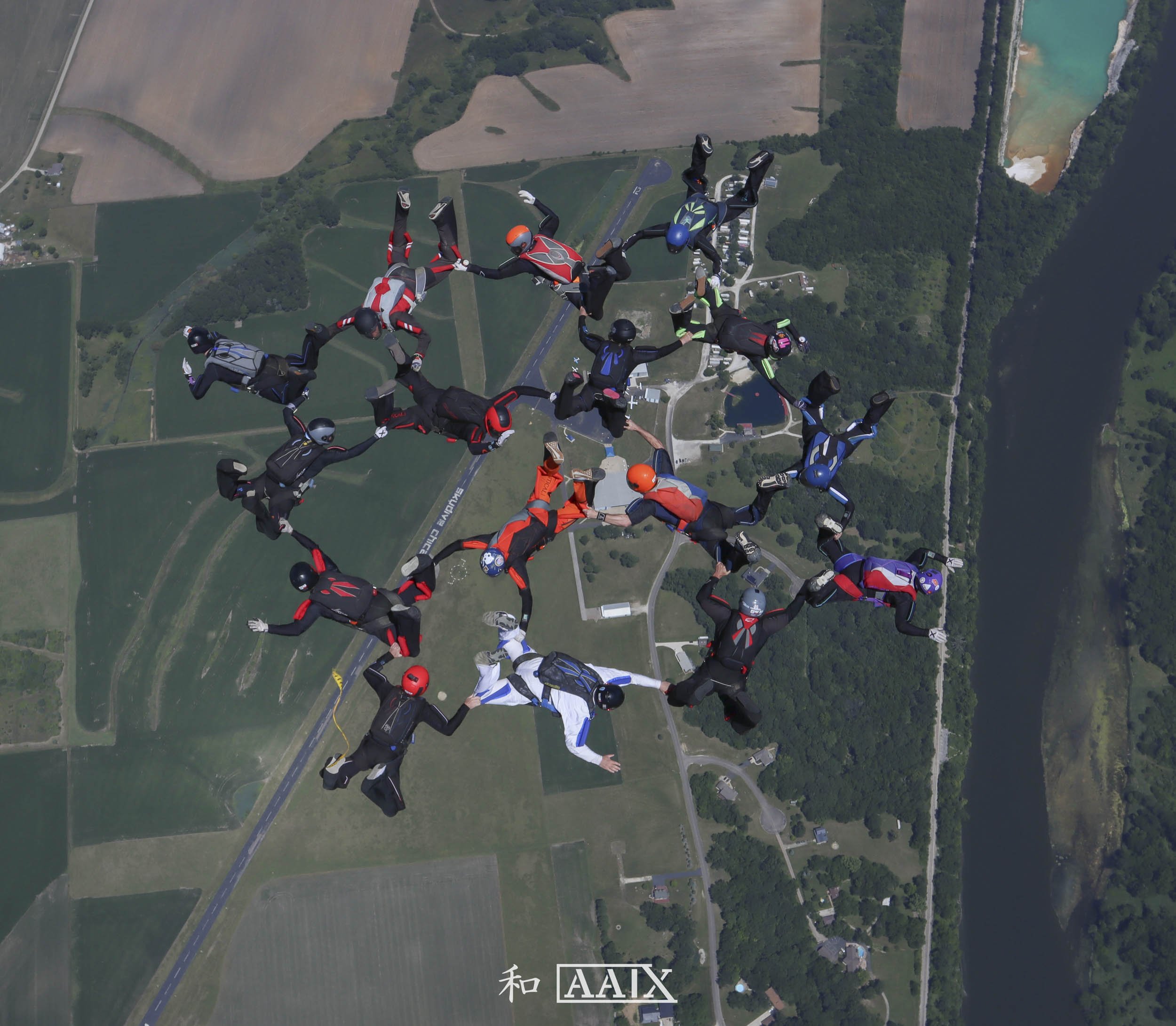 Group of skydivers in freefall holding hands over a rural landscape with fields, trees, and a river.