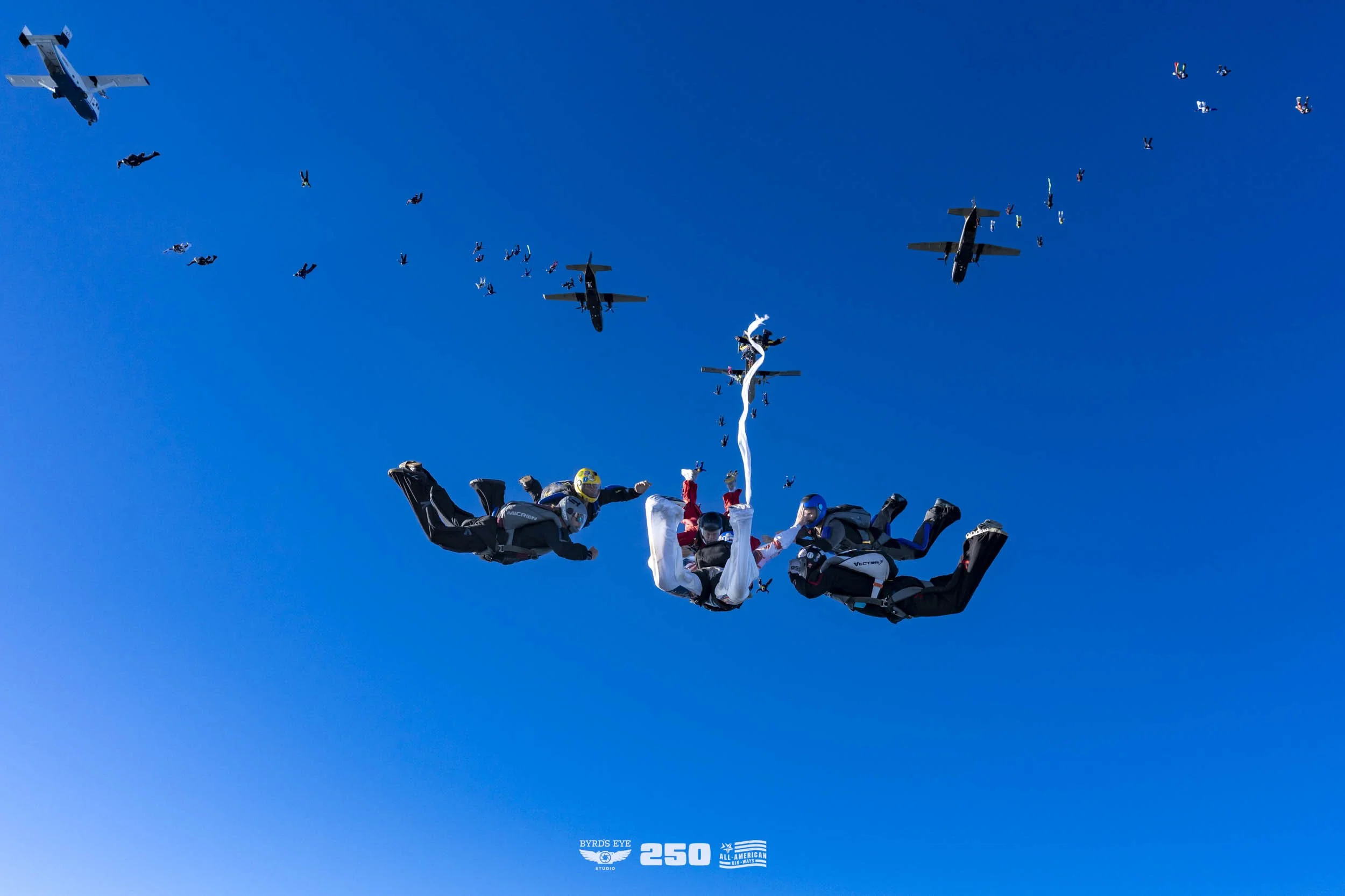 Skydivers in freefall formation with airplanes flying in a clear blue sky.