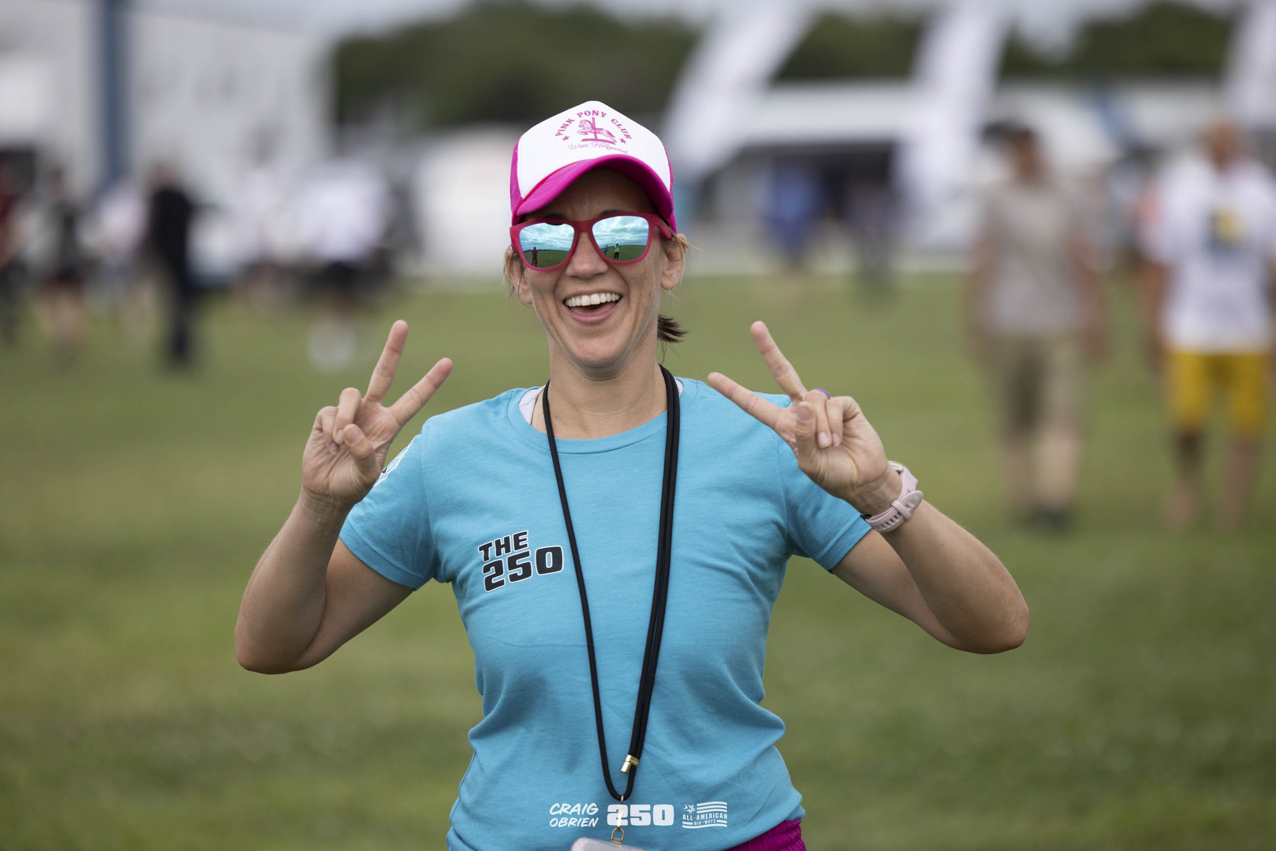 Woman smiling and making peace signs with both hands at a running event, wearing sunglasses, a pink and white cap with a pony club logo, a blue t-shirt with event details, and a black lanyard, outdoors with other participants and vehicles in the back