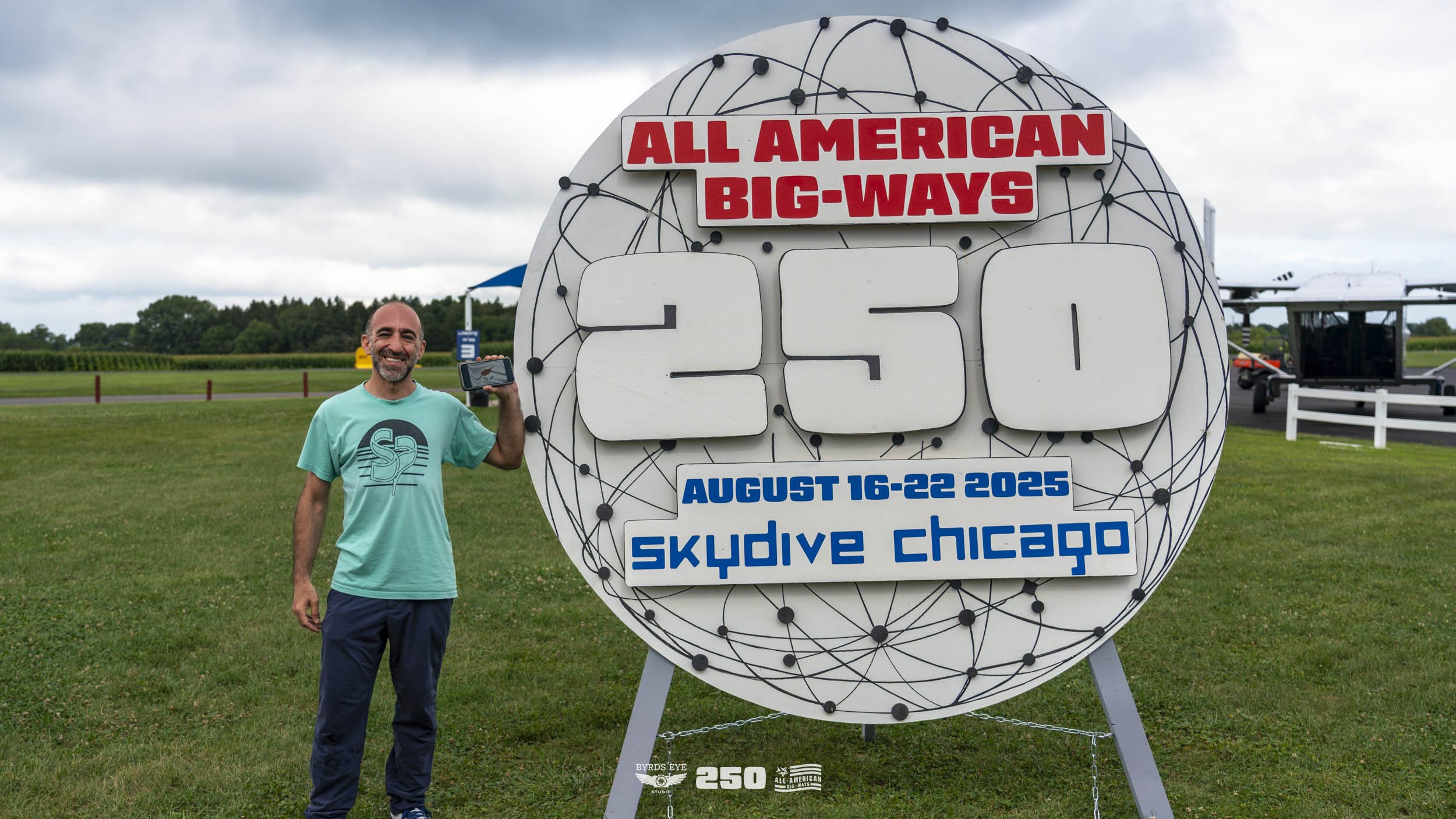 A smiling man standing next to a large sign that reads 'All American Big-Ways 250' with dates August 16-22, 2025, for Skydive Chicago. The man is holding a phone, standing on a grassy field with open skies, a small aircraft, and trees in the backgrou
