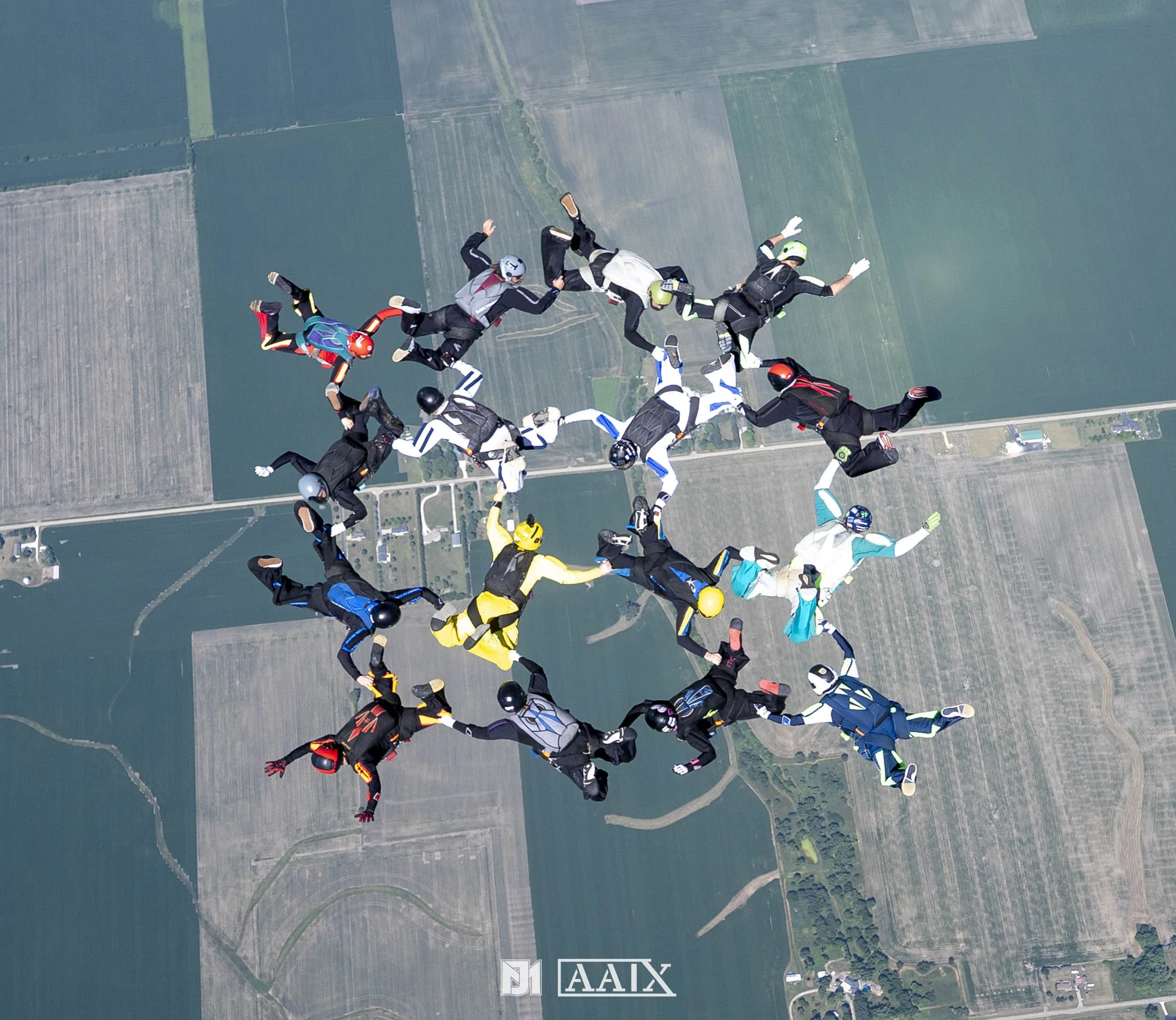 A group of skydivers in jumpsuits and helmets forming a star shape while free-falling above farmland.