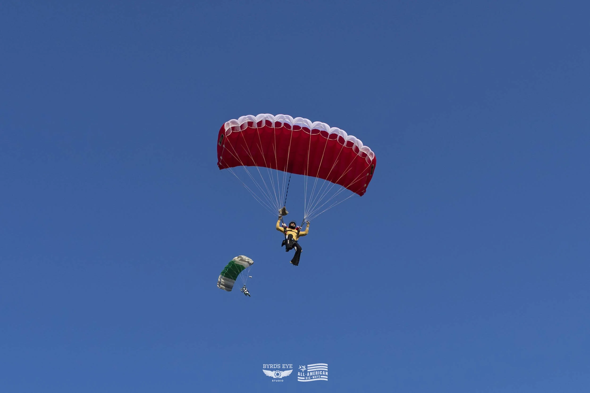A person skydiving with a red and white canopy in a clear blue sky, with a smaller green and white canopy in the background.