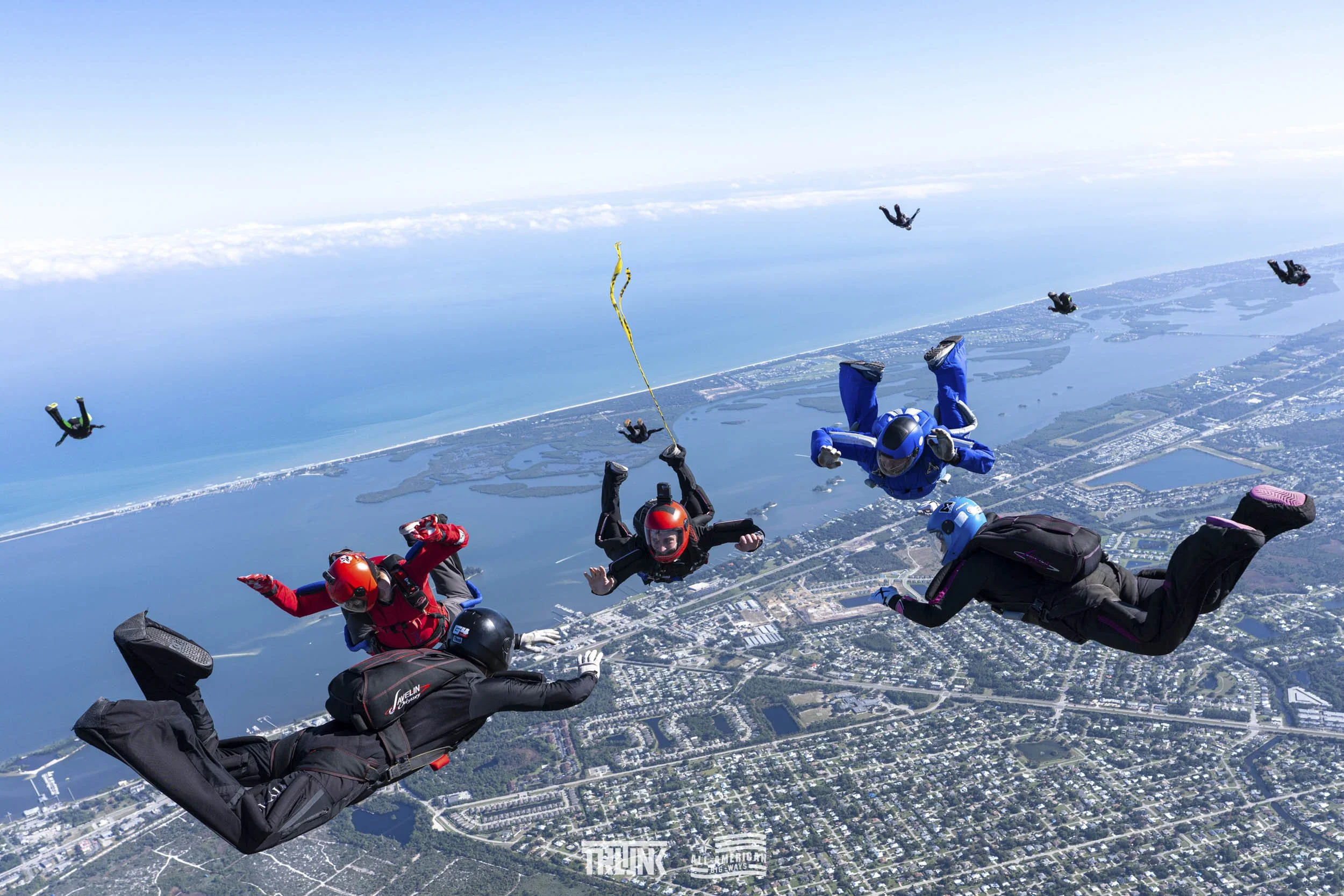 Skydivers in free fall over a coastal city with water, land, and roads visible below.