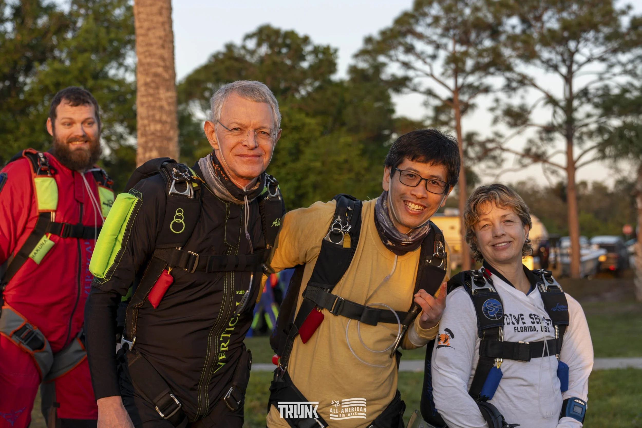 Four skydivers prepare for a jump, smiling and posing outdoors during sunset, with trees and parked cars in the background.