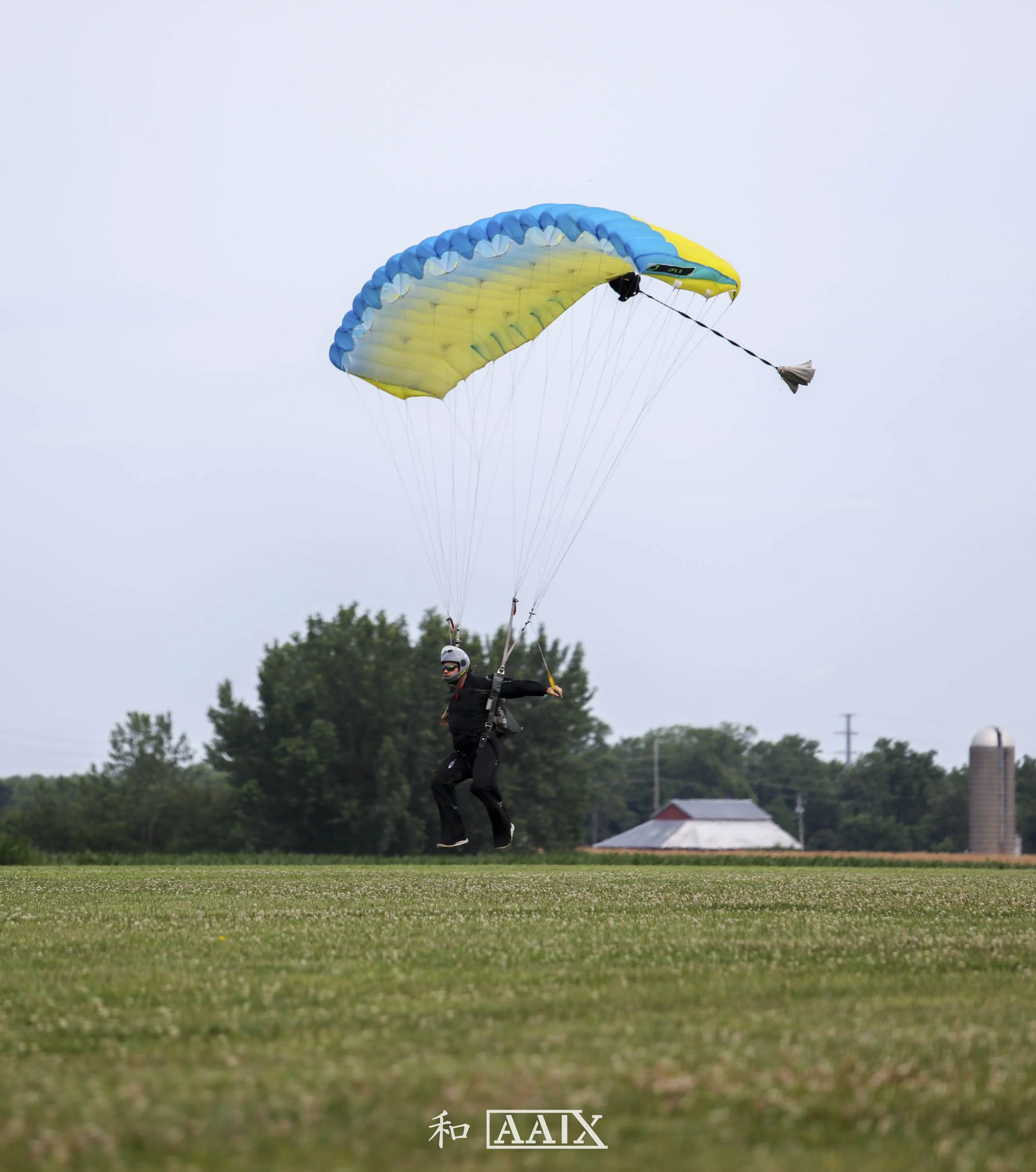 Person with a parachute on a grassy field, preparing for or landing from a parachute jump, overcast sky and farm buildings in the background.