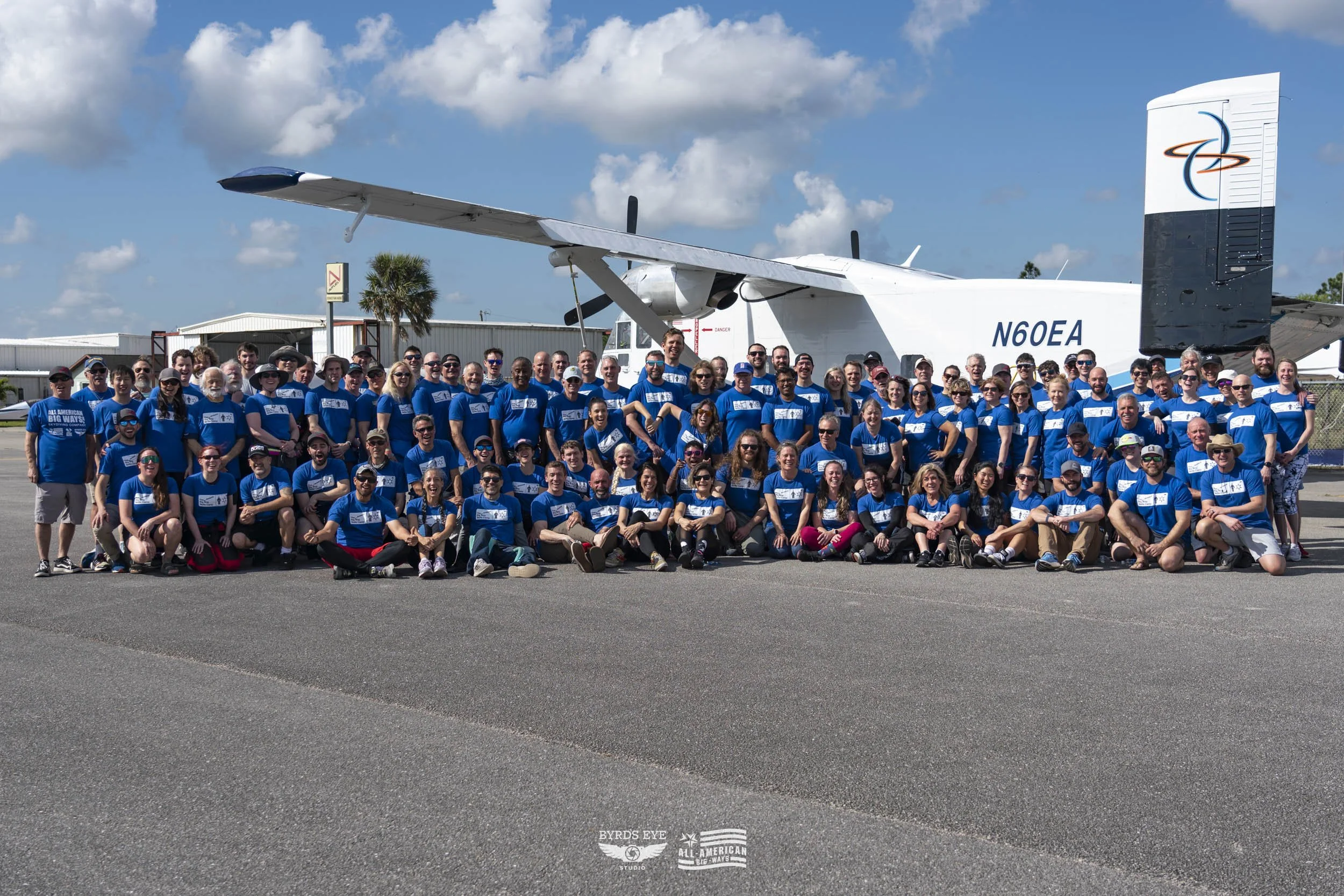 Group of people in blue shirts posing in front of a large white aircraft on an airfield, under a partly cloudy sky.