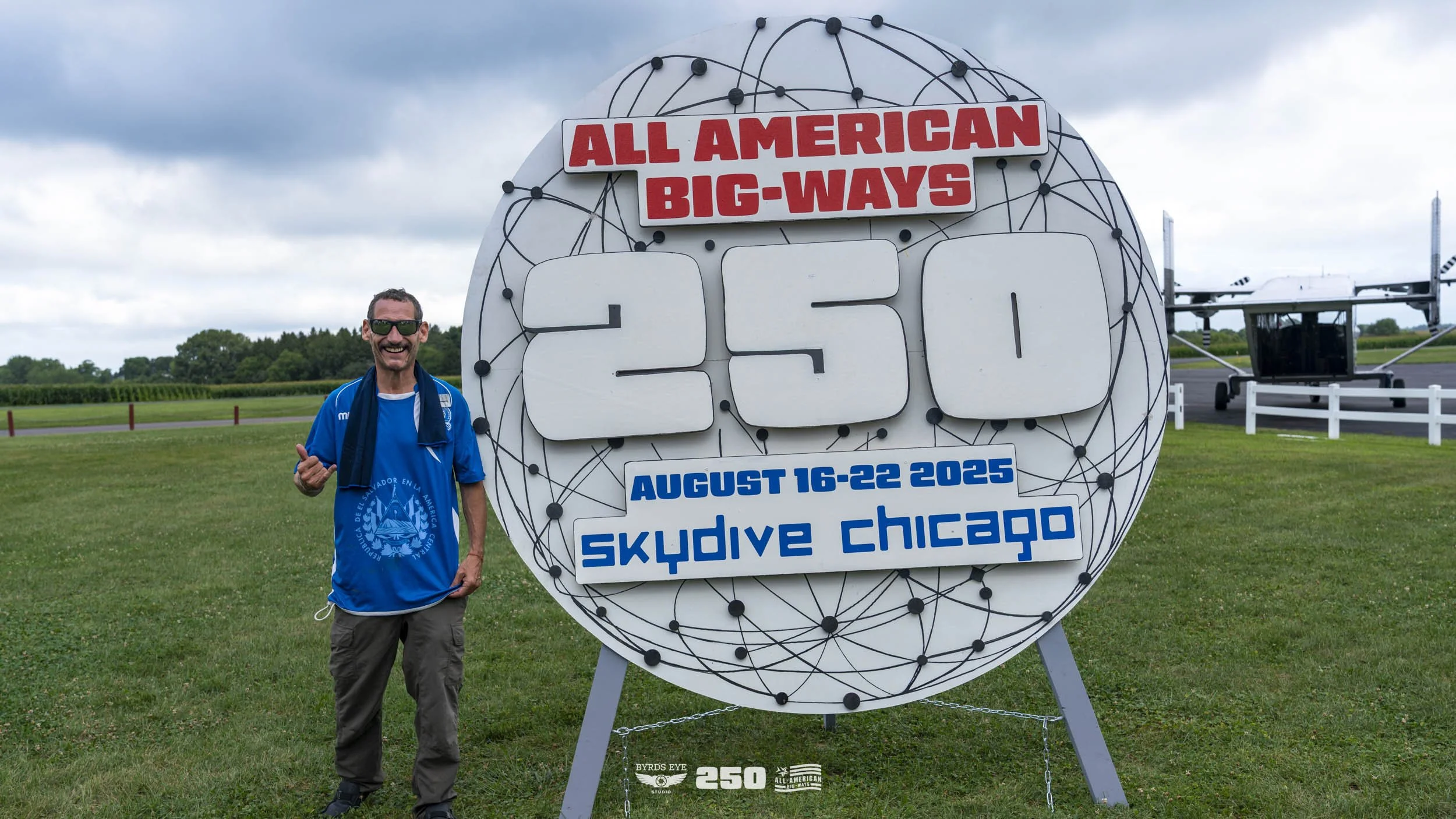 A smiling man stands next to a large sign that reads "All American Big-Ways 250" with details about a skydiving event in Chicago from August 16-22, 2025. The man is wearing a blue shirt and sunglasses, standing on green grass with open farmland and a