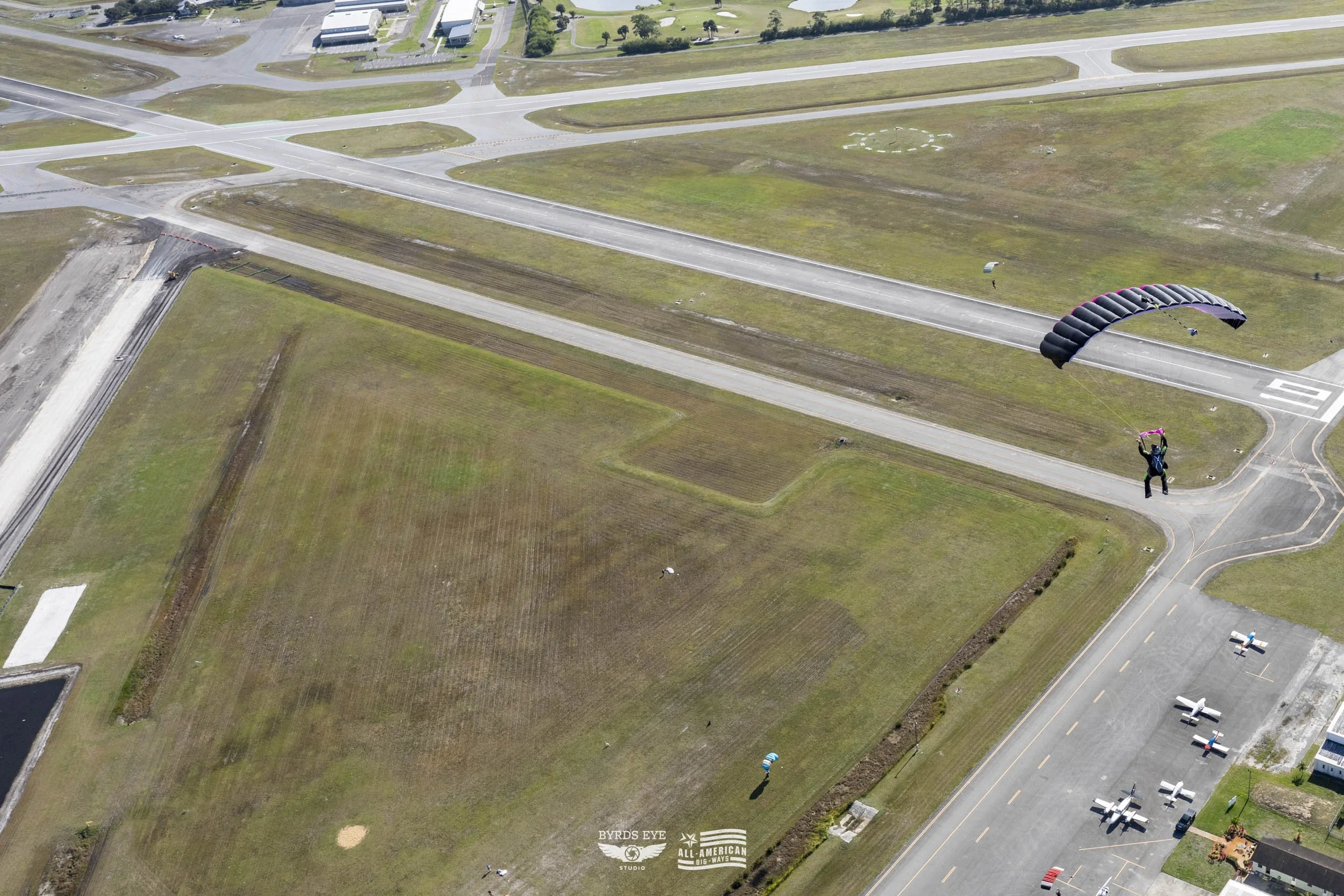 A person skydiving with a black parachute over an airfield with fields, roads, and parked aircraft.