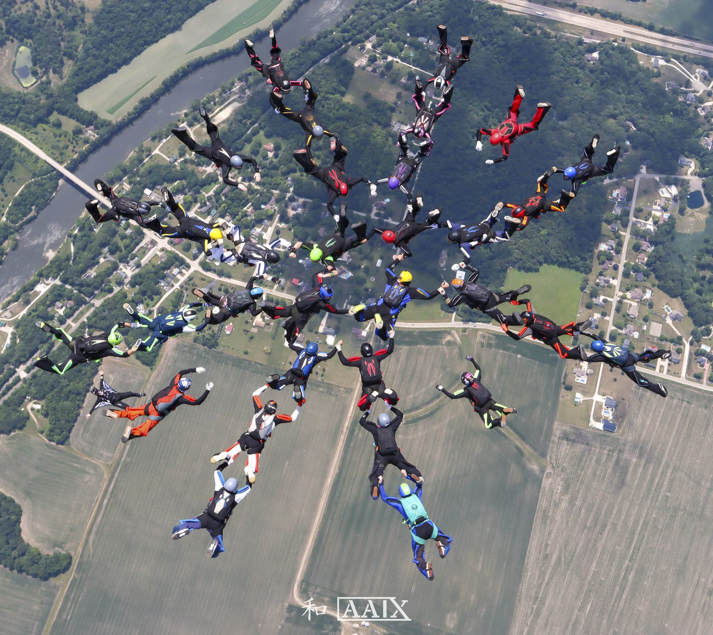 Group skydivers in freefall formation over a rural landscape with fields, roads, and a river.