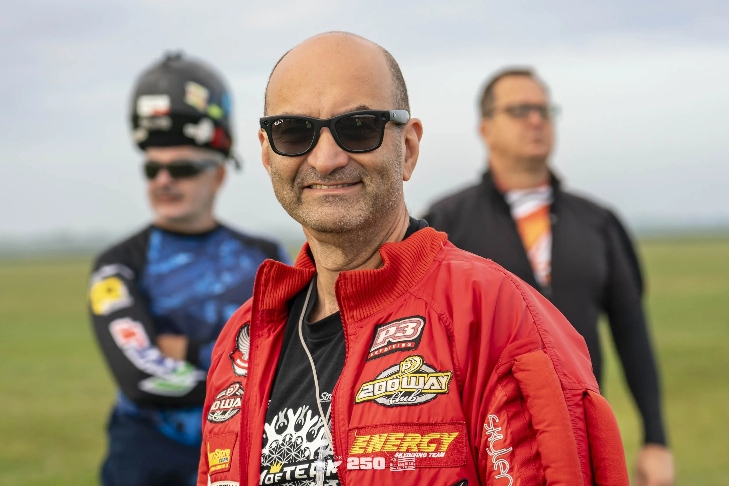 A smiling man with bald head and sunglasses in the foreground, wearing a red racing jacket with various patches and logos. Behind him, two men wearing glasses and racing gear stand outdoors on a grassy field, with a cloudy sky overhead.