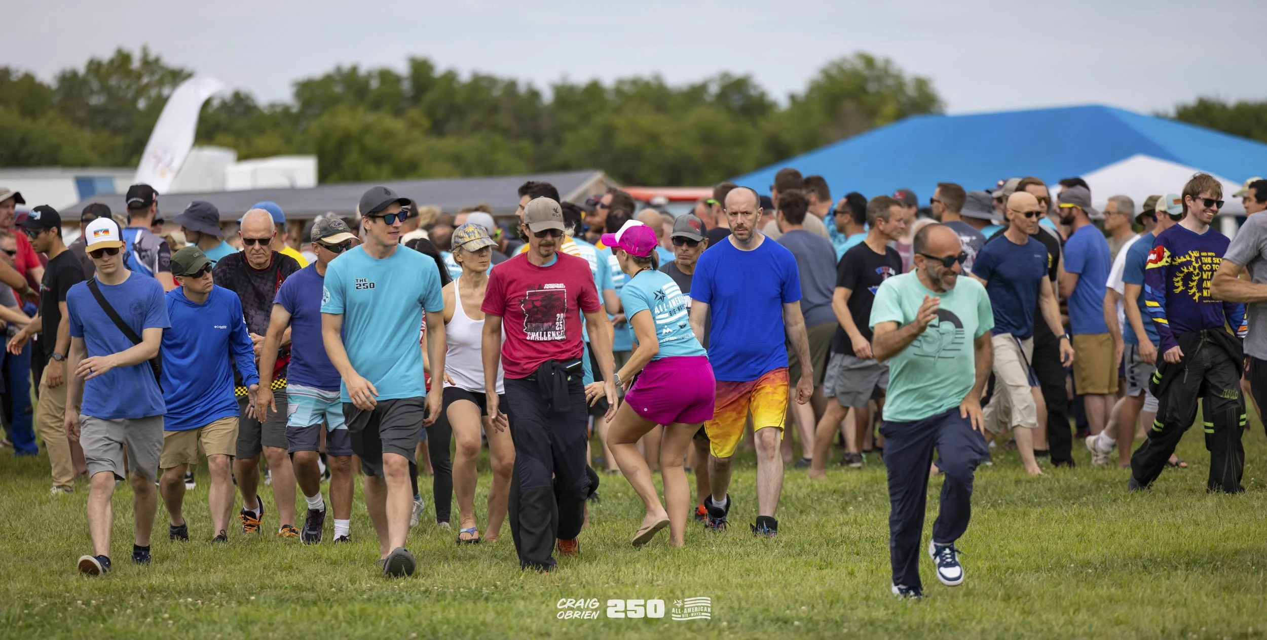 A large group of people walking on a grassy field during an outdoor event, with some wearing casual clothes, sunglasses, and hats, and a few in athletic apparel.