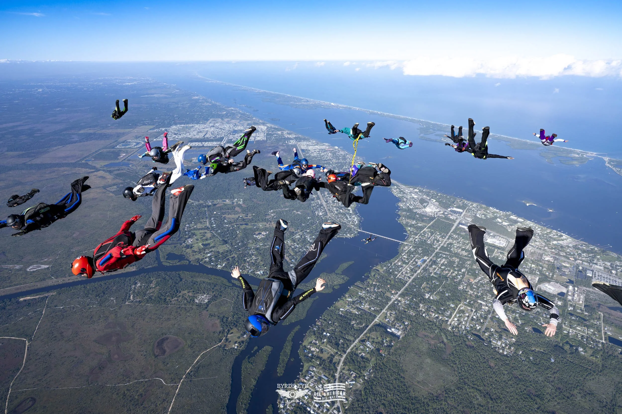 Group of skydivers in freefall over a landscape with water, trees, and buildings, under clear blue sky.