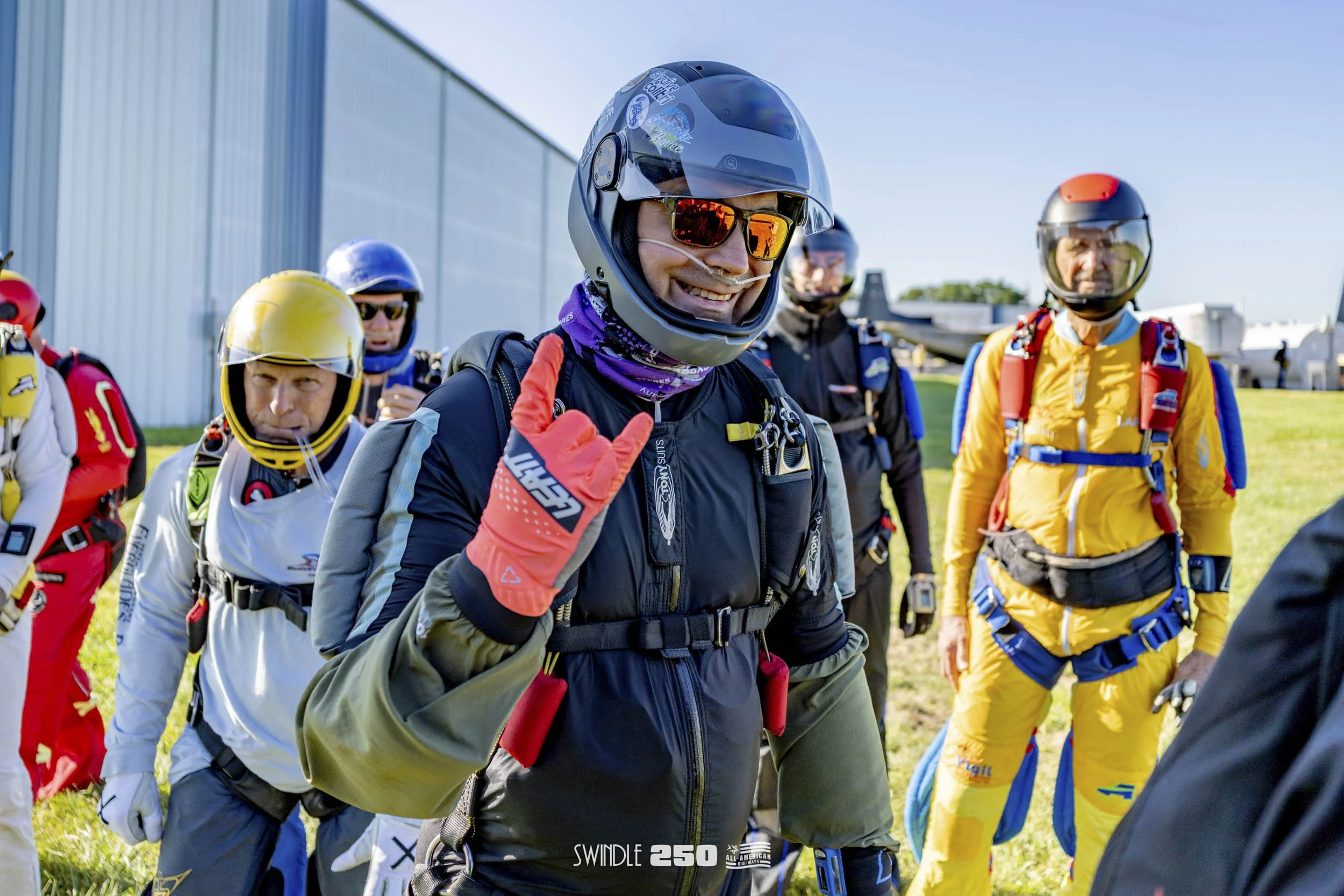 Group of skydivers in colorful jumpsuits and helmets standing outdoors on a grassy field, preparing to jump with an airplane in the background.