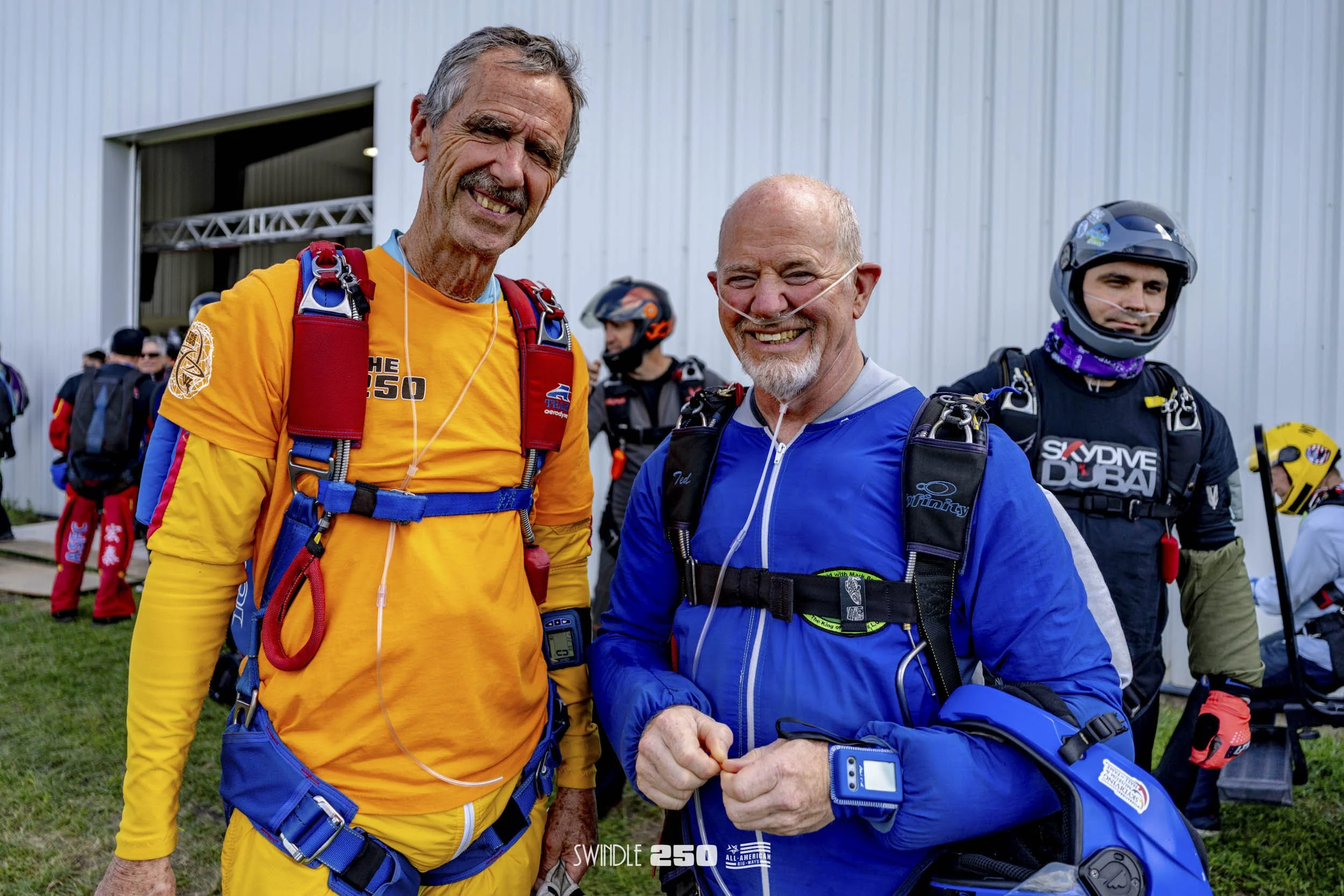 Two elderly men smiling, wearing skydiving gear, standing outside a hangar with other skydivers in the background.