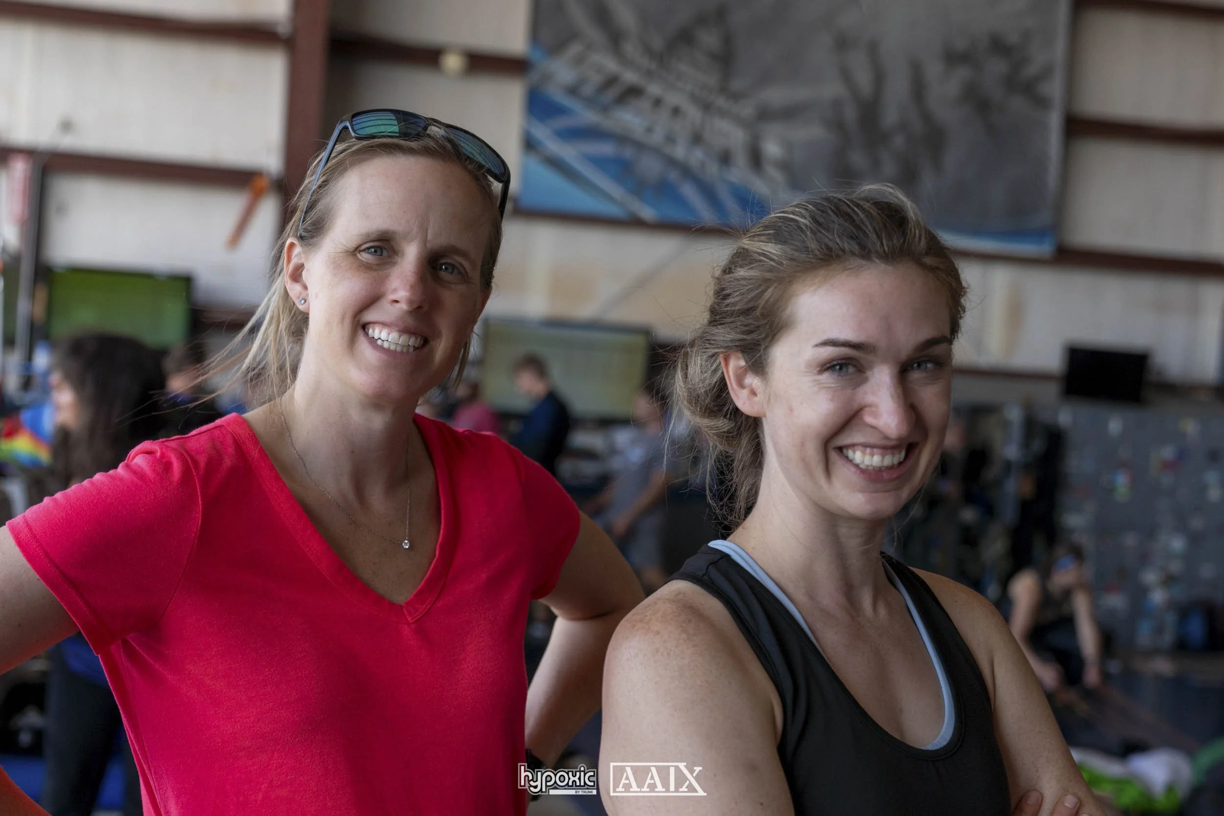 Two women in workout attire smiling inside a gym or training facility with other people working out in the background.