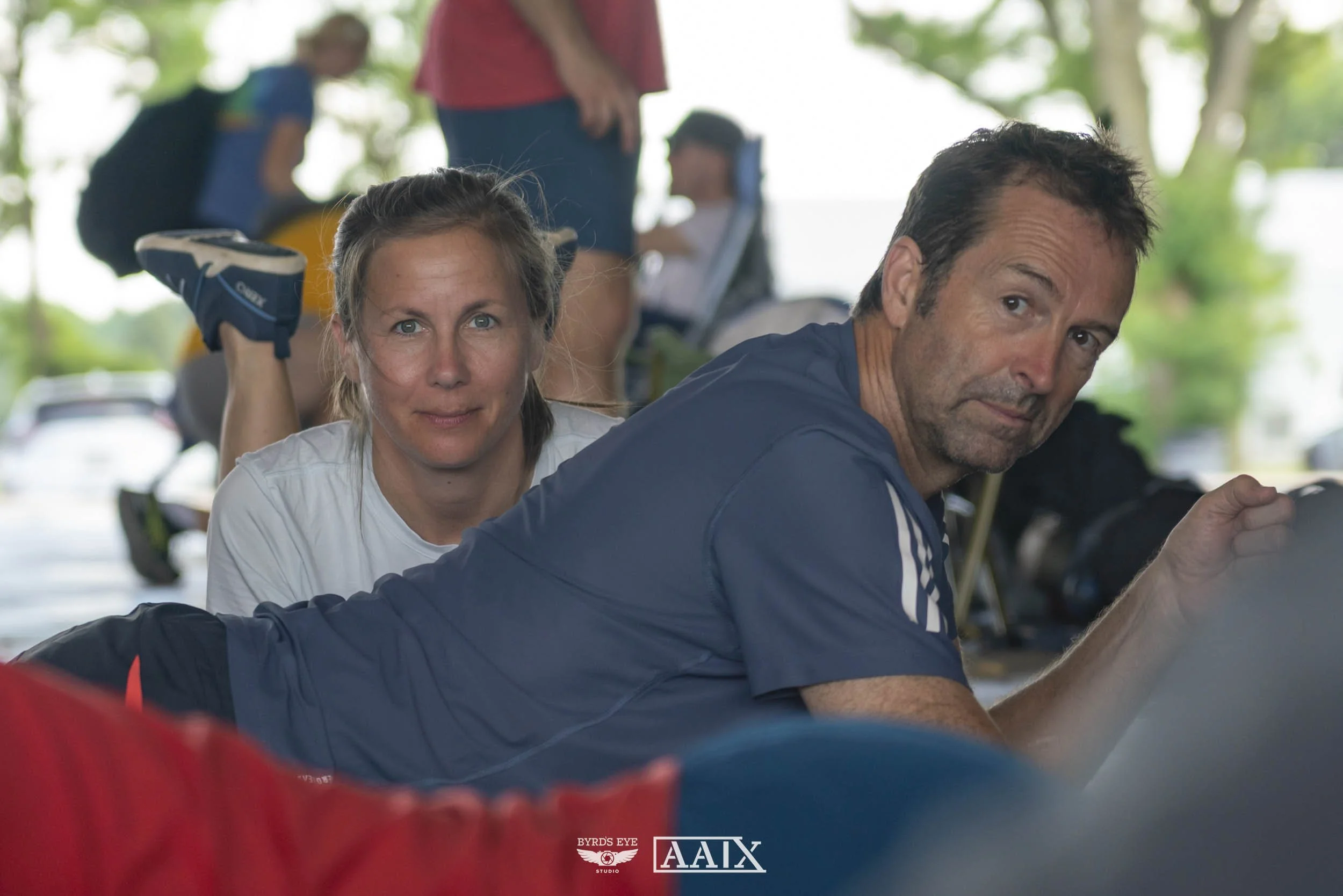 A man and a woman are lying on the ground in a gym or training area, with the woman looking directly at the camera and the man slightly turned away, both appearing to be doing some kind of exercise. In the background, there are children and a woman s